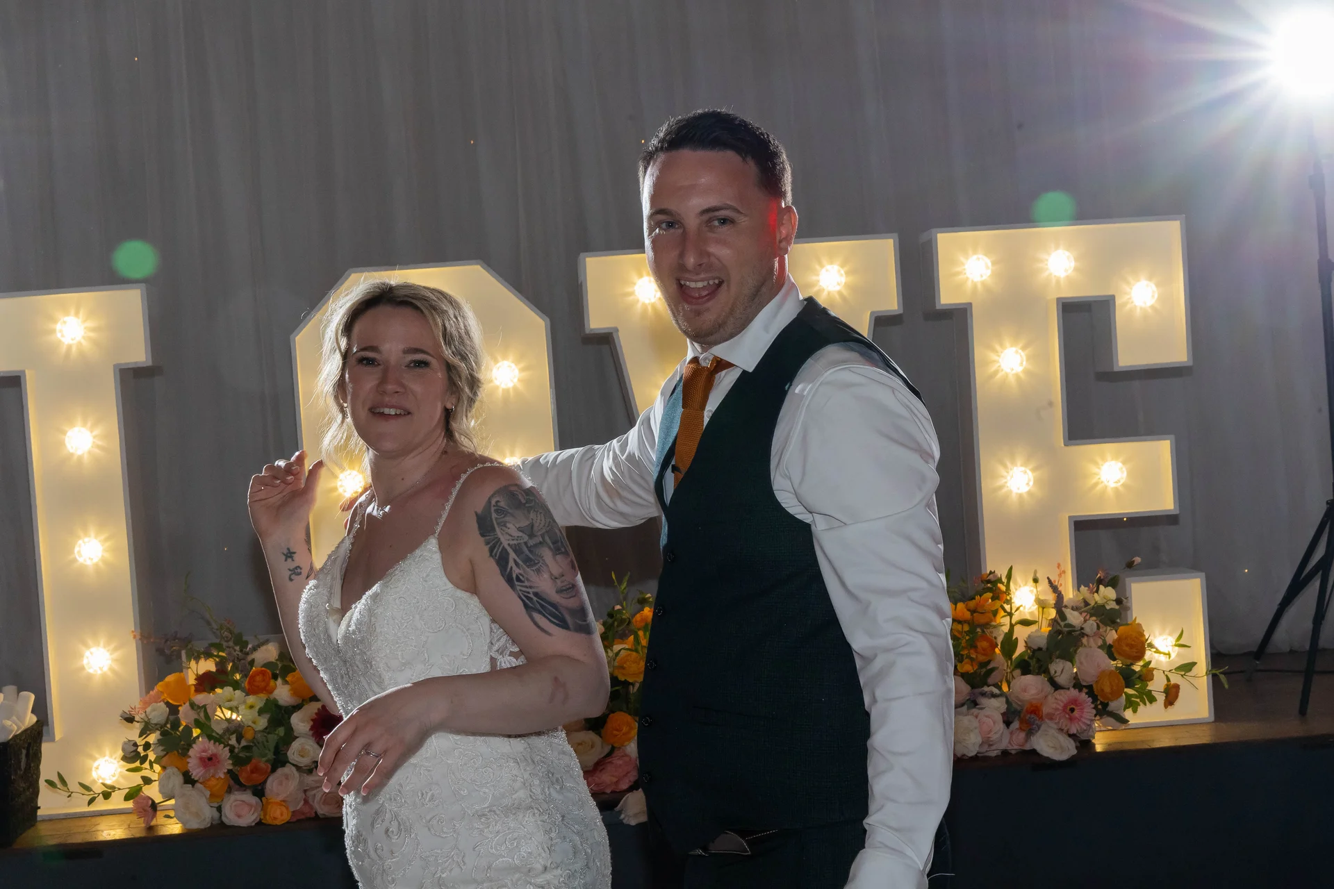 A newlywed couple laugh and dance together on the dance floor in front of illuminated giant love letters at their evening reception.
