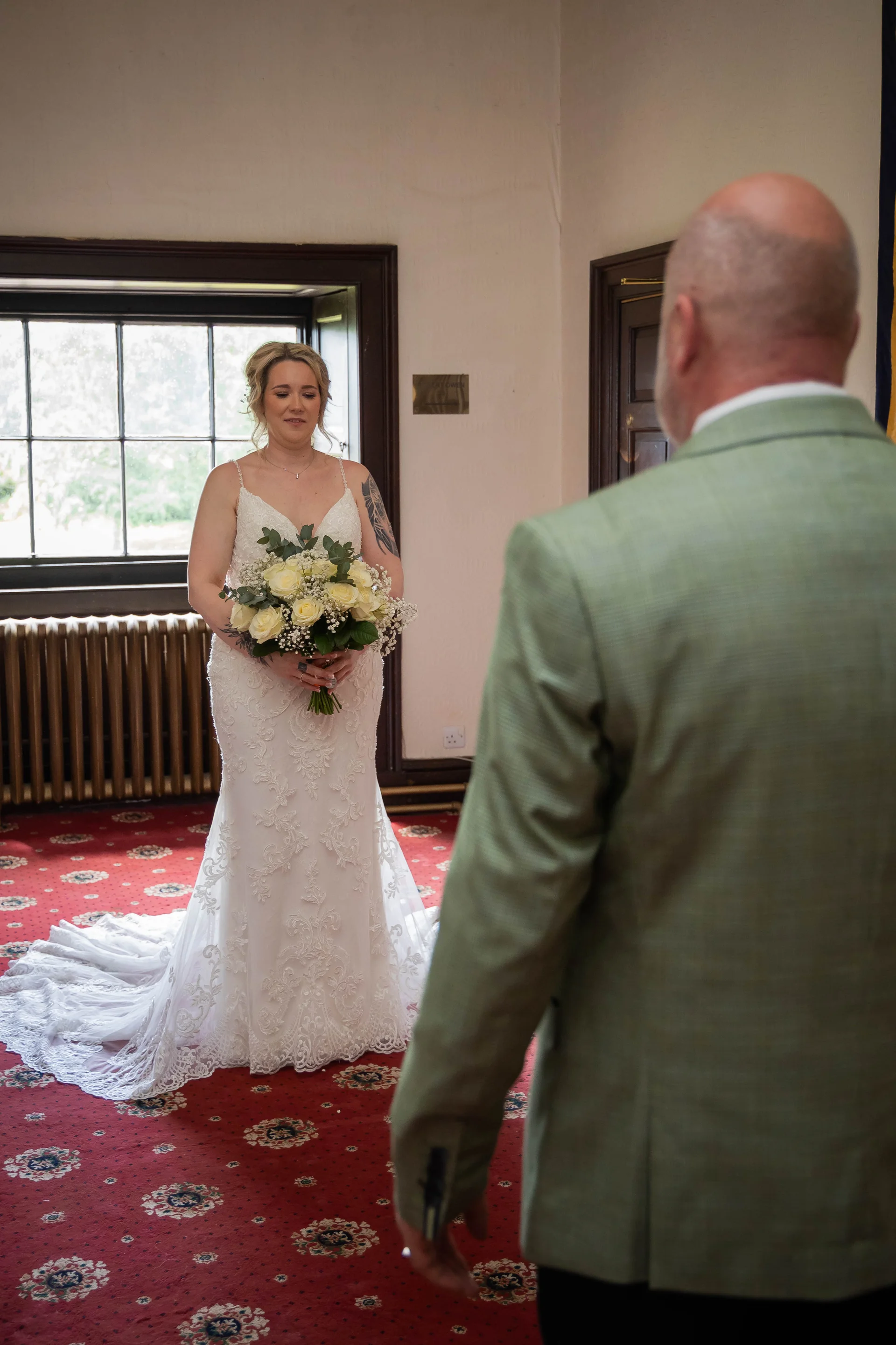 A bride in a lace gown with a bouquet of white roses shares a tender first look moment indoors with a family member wearing a green suit.