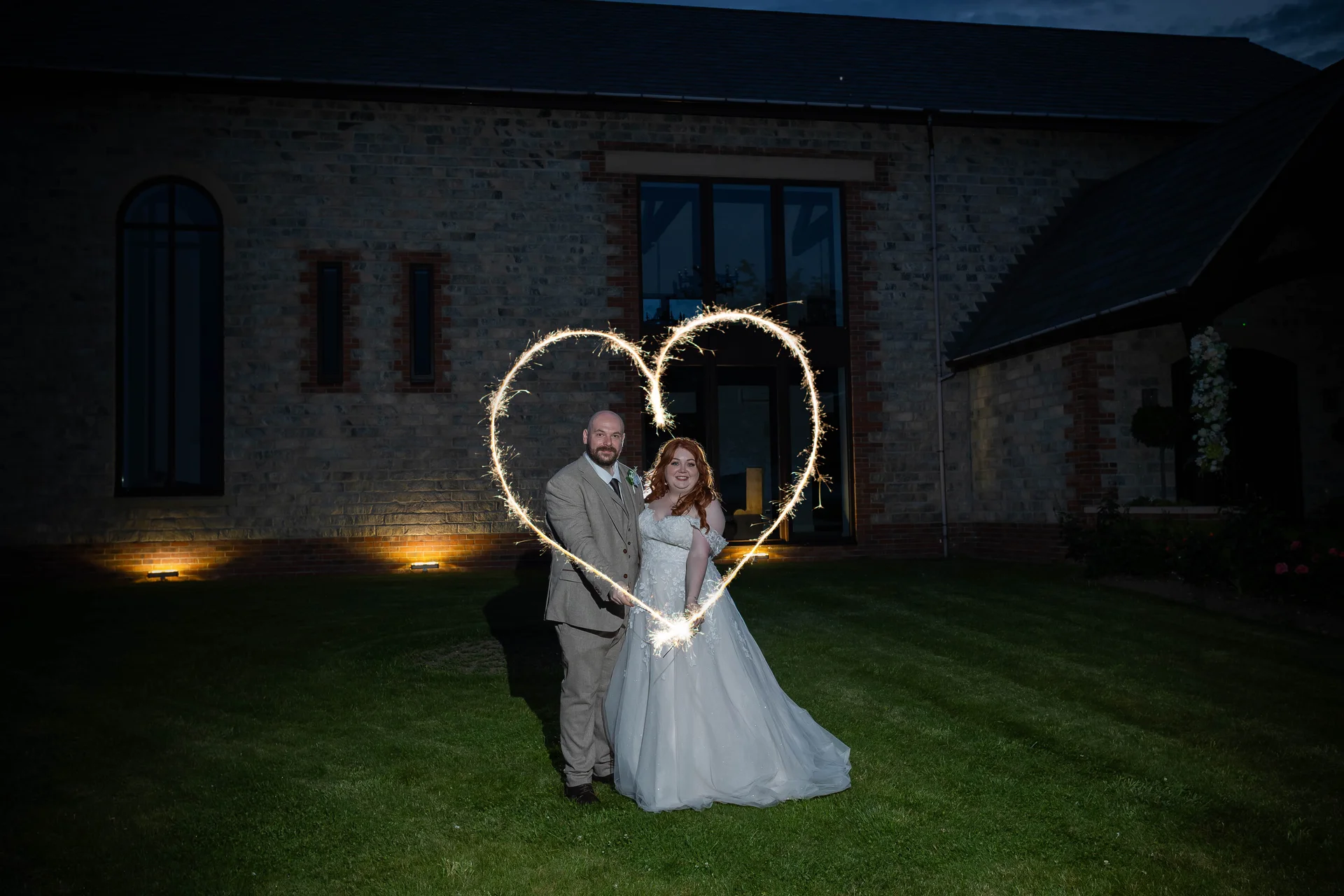 A newlywed couple poses in front of a stone barn at night while creating a glowing heart shape with sparklers.