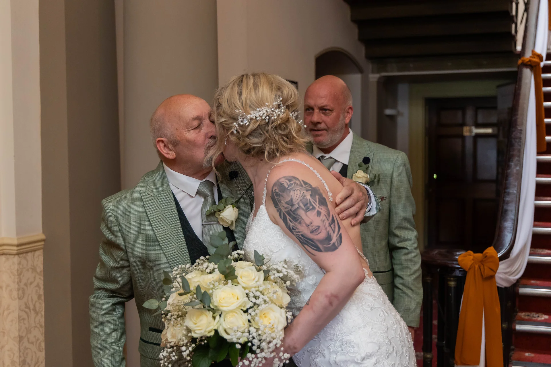 A father affectionately kisses his bride daughter on the cheek as she holds a large bouquet of white roses indoors near a grand staircase.