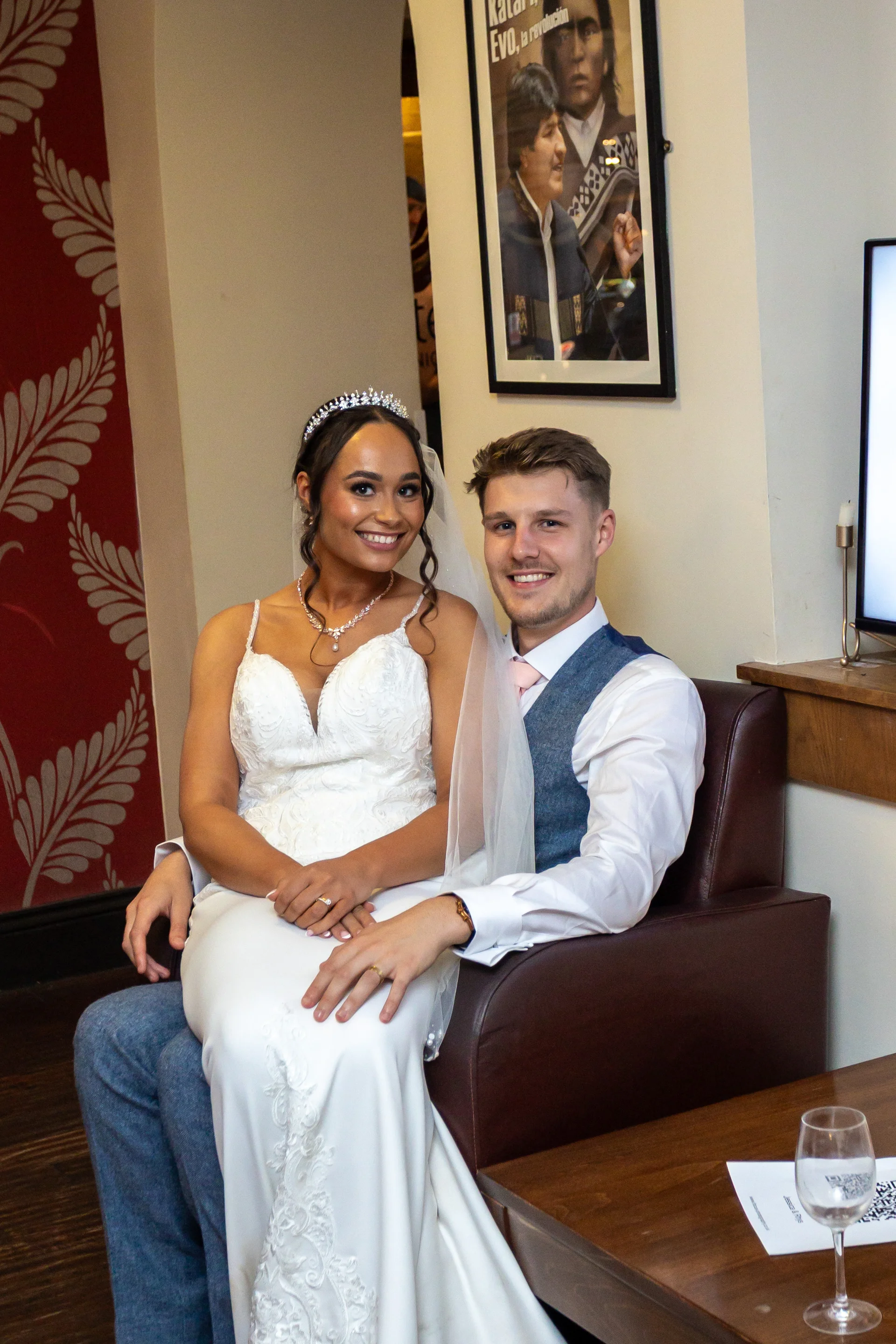 A smiling bride wearing a beaded tiara and lace-detailing gown sits on her groom's lap in a cosy interior setting.