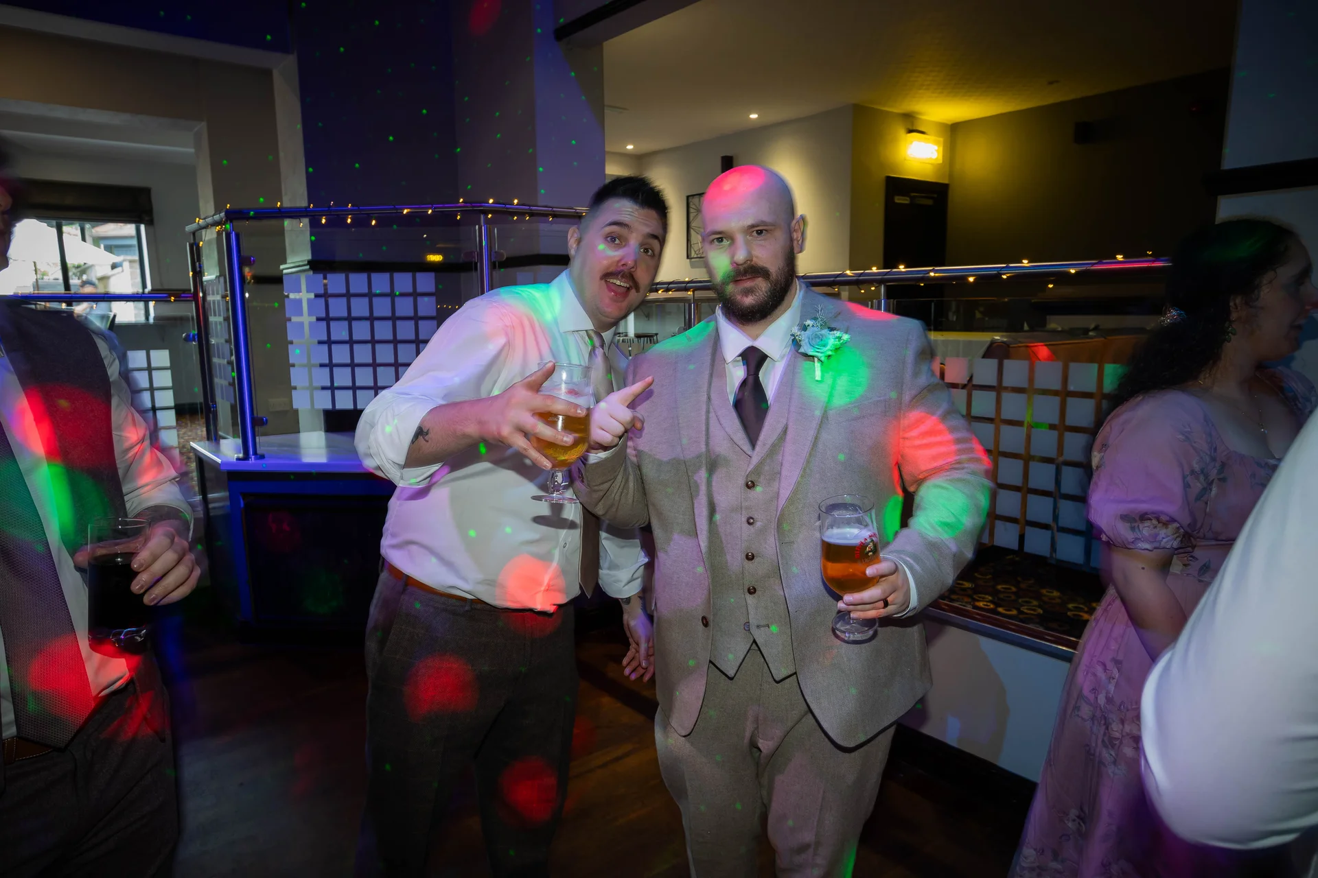 Two wedding guests enjoy a drink and pose for a photo on the dance floor under colourful disco lights.