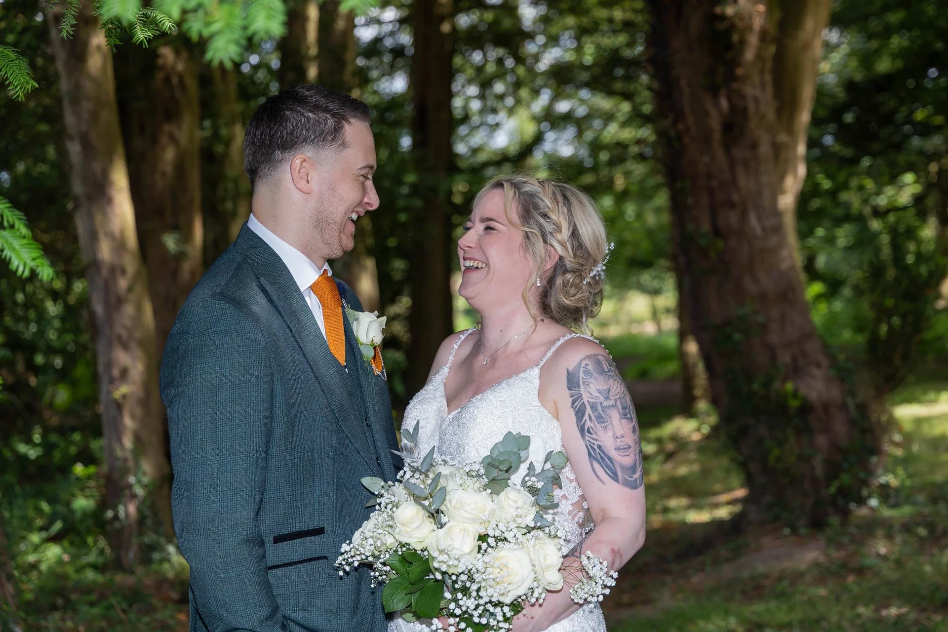 A newlywed couple shares a joyful belly laugh while standing outdoors in a lush woodland setting surrounded by trees.