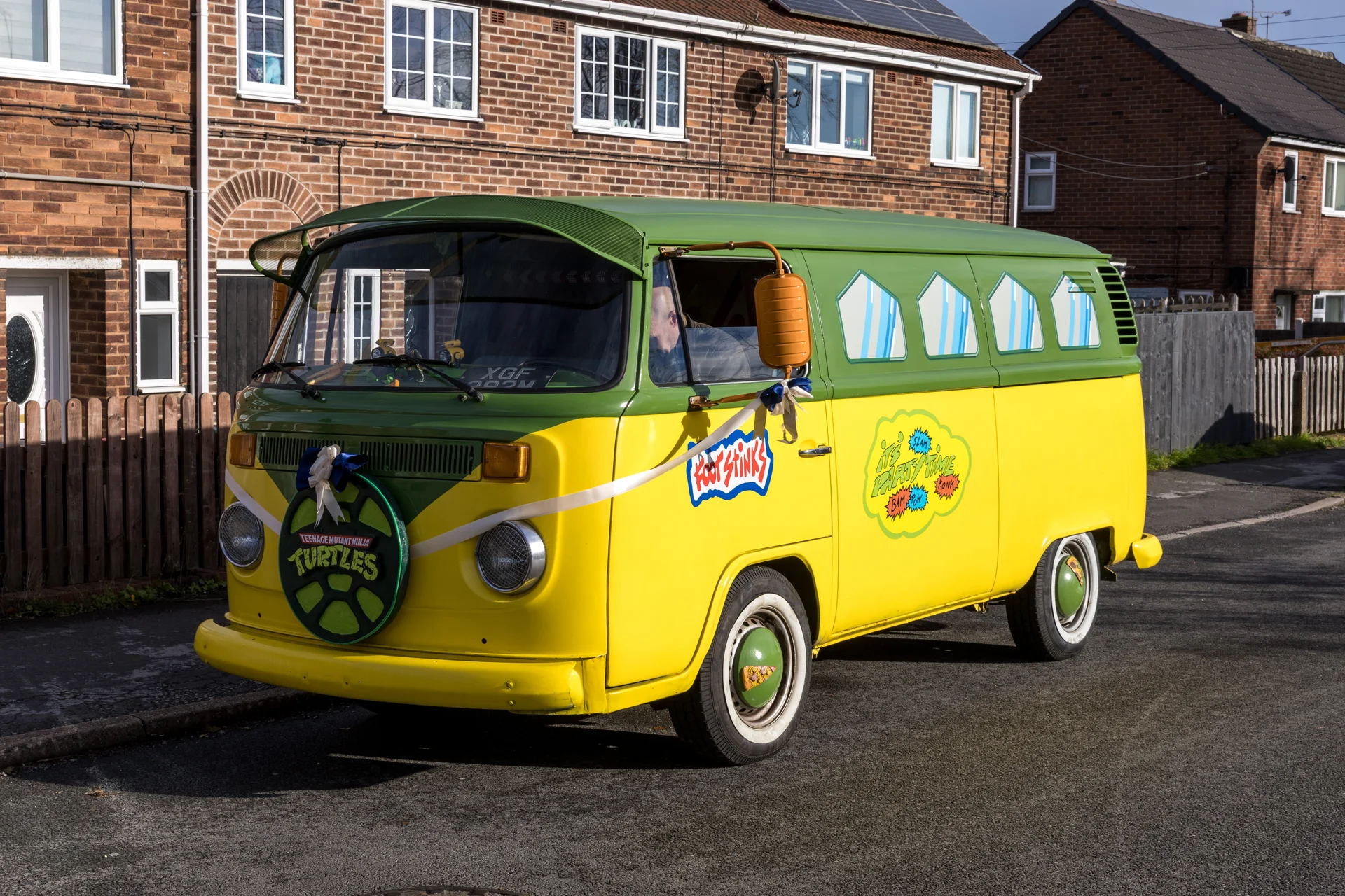 A vibrant yellow and green vintage Volkswagen campervan decorated for a wedding with ribbons and a Teenage Mutant Ninja Turtles theme is parked on a suburban street.