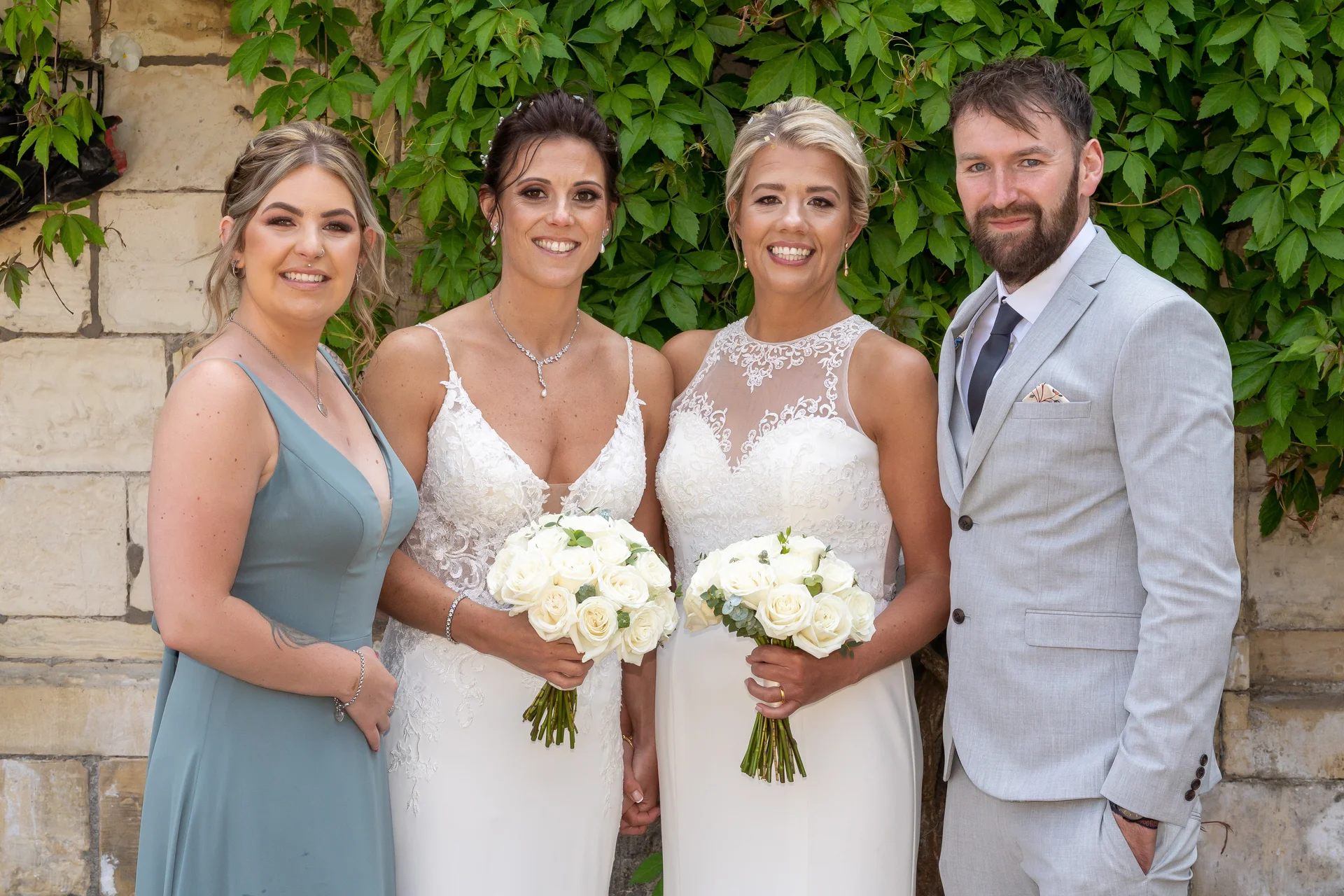 A smiling wedding party featuring two brides in white lace gowns holding rose bouquets, alongside a bridesmaid in sage green and a groom in a light grey suit against an ivy-covered stone wall.