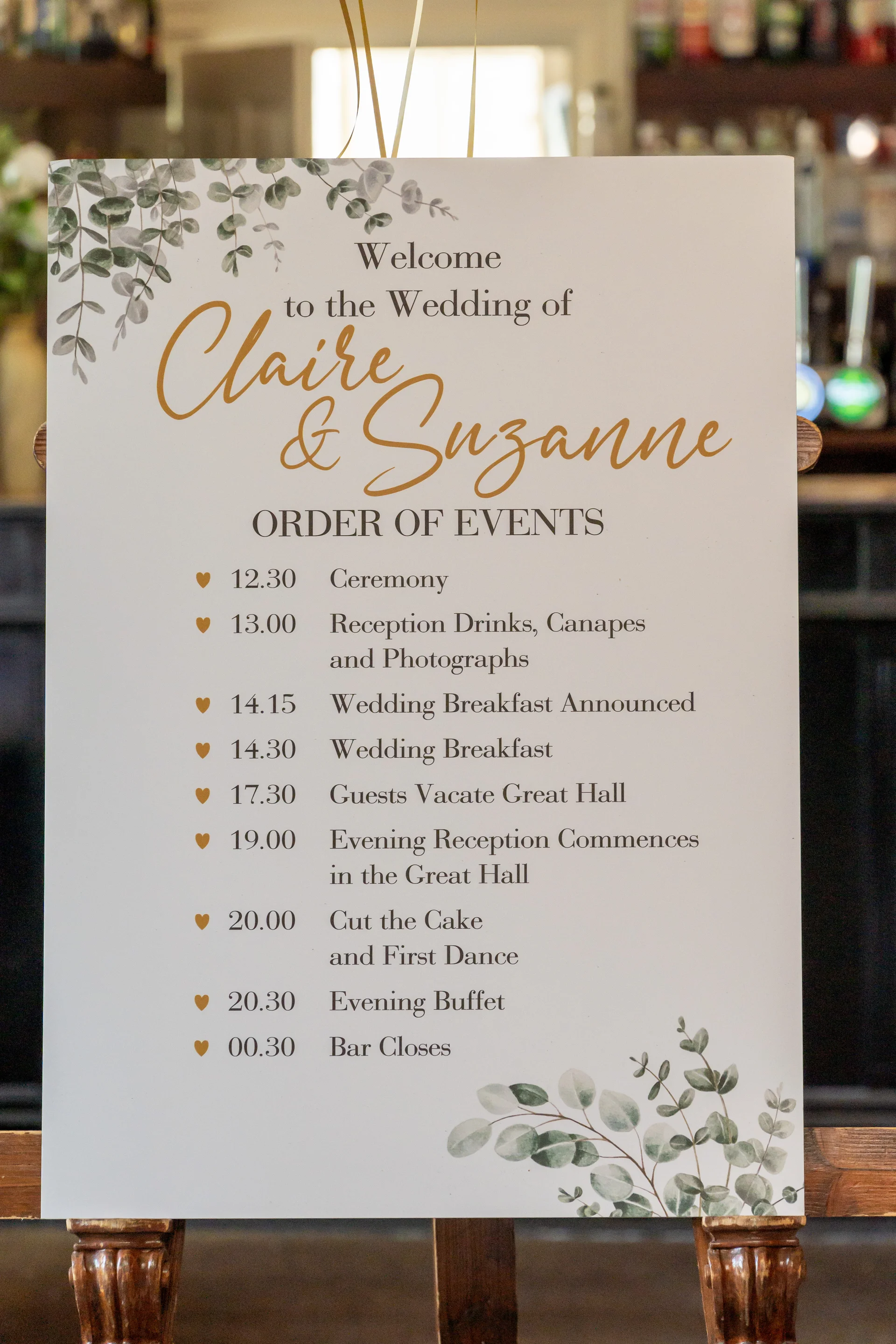 An elegant wedding welcome sign and order of events displayed on a wooden easel, featuring eucalyptus foliage illustrations and gold script lettering for two brides.