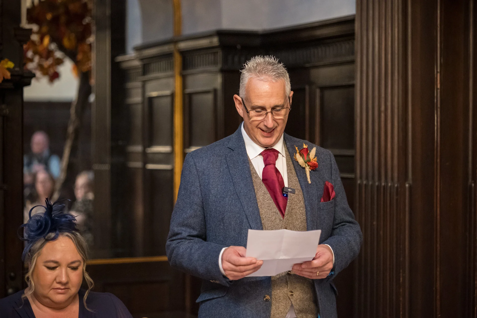A smiling groom wearing a blue tweed suit and brown waistcoat delivers a wedding speech while holding notes in a wood-panelled room.