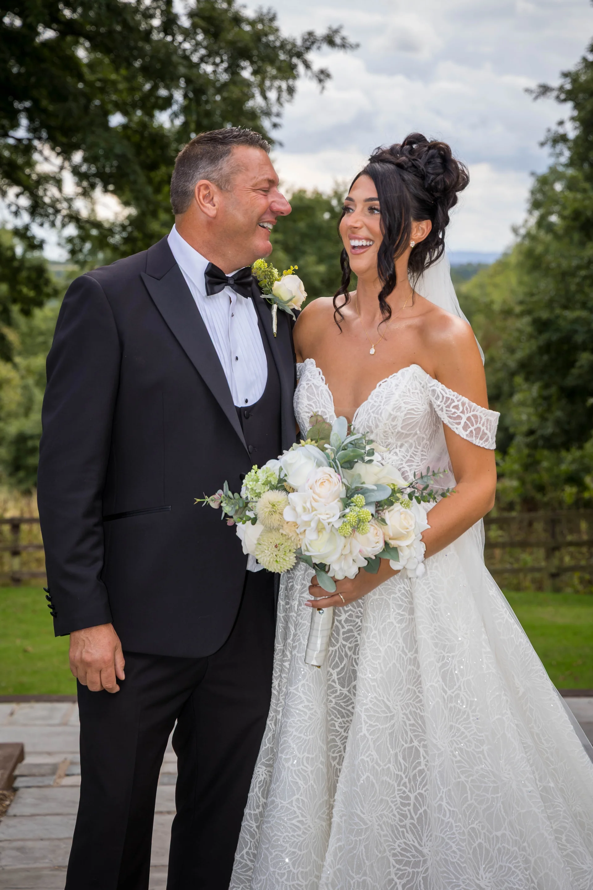 A newlywed couple shares a joyful laughing moment outdoors, with the bride holding a lush white floral bouquet and wearing a lace gown.