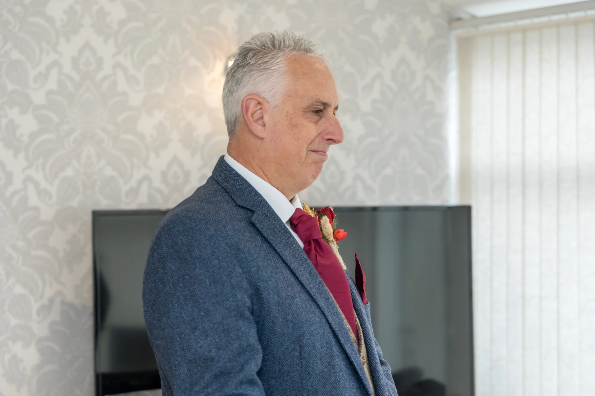 A profile portrait of a groom wearing a blue textured suit and a burgundy tie with a floral buttonhole.