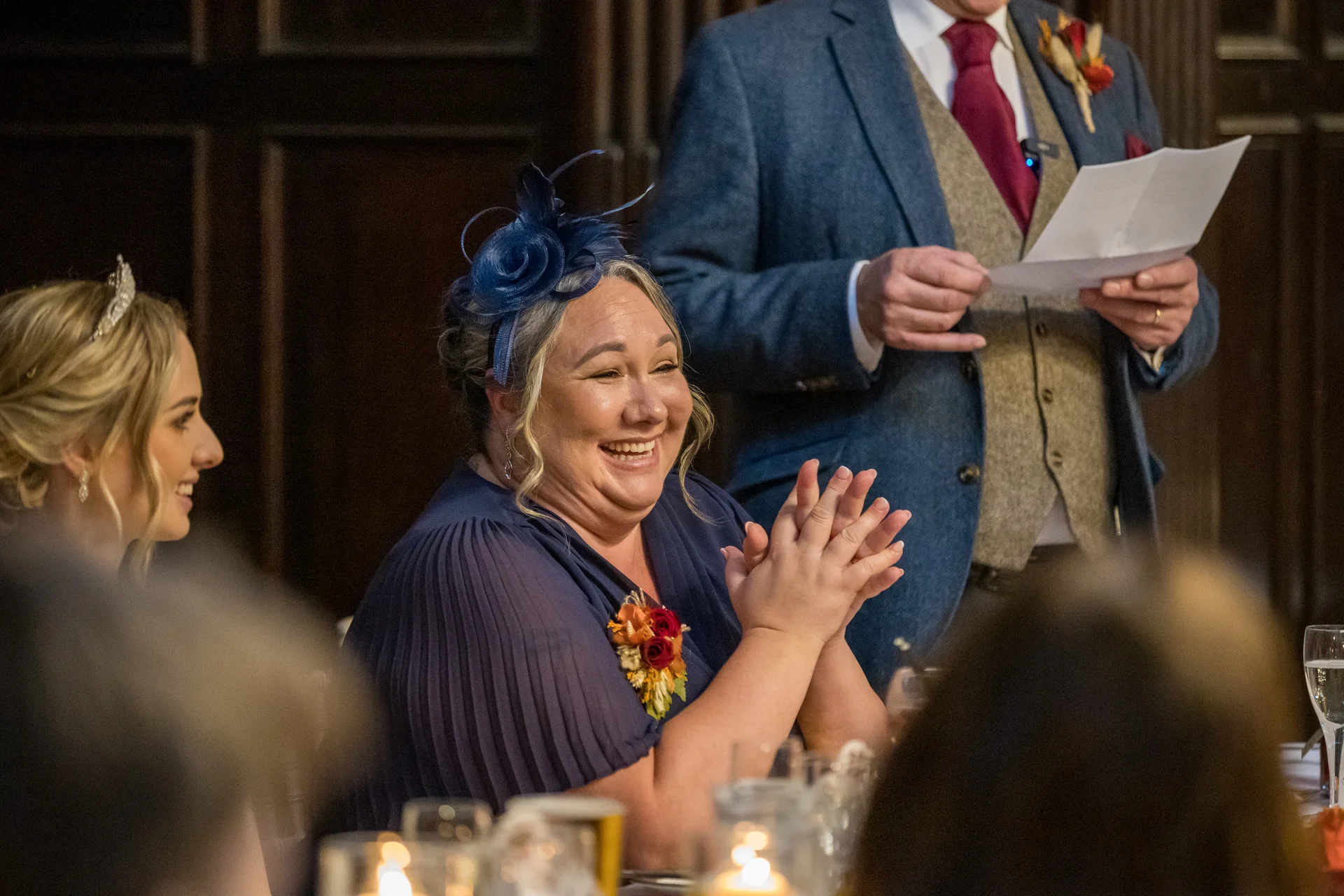 A guest wearing a blue fascinator and navy dress claps and laughs happily during the wedding speeches while another guest reads notes nearby.