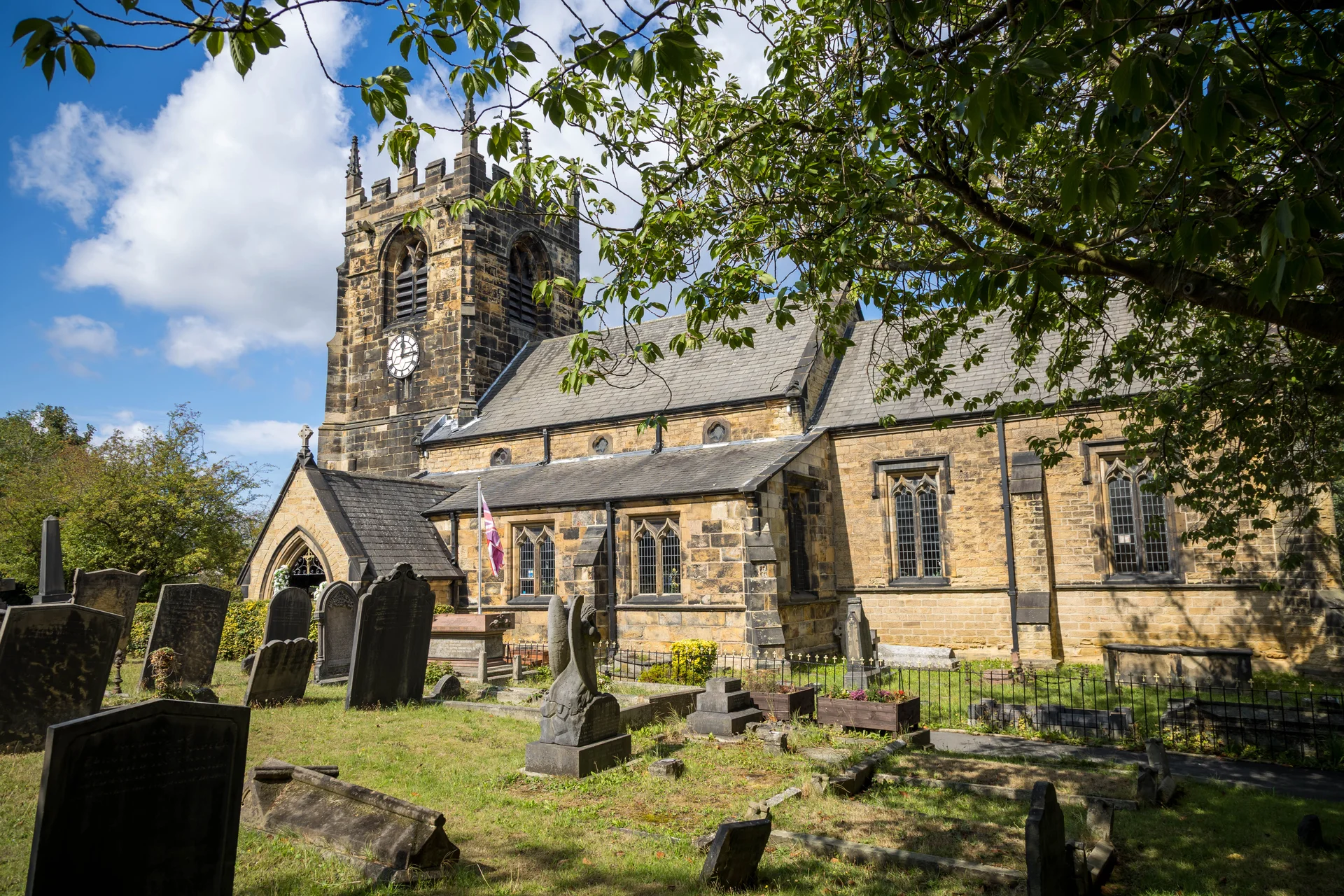 A historic stone church with a clock tower and graveyard stands under a bright blue sky on a sunny day, viewed through overhead tree foliage.