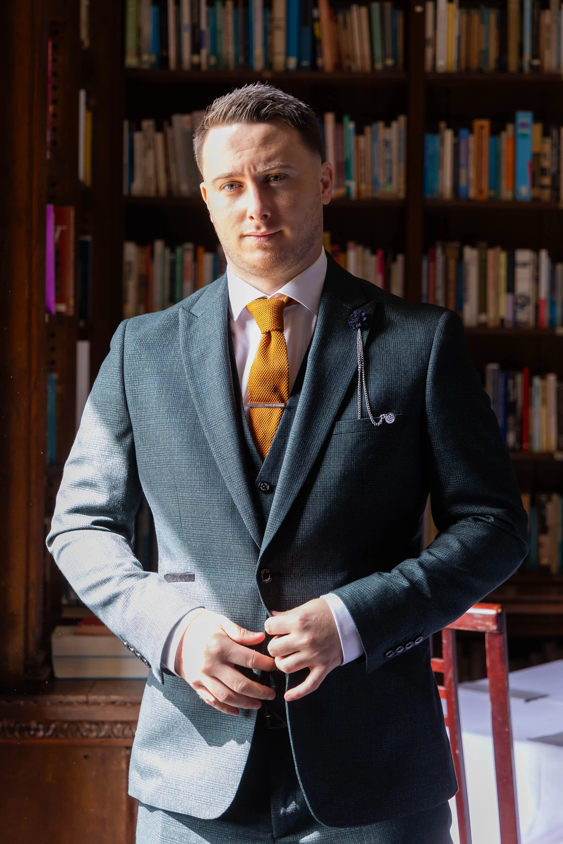 A handsome groom in a textured dark grey three-piece suit and burnt orange tie poses confidently inside a library.