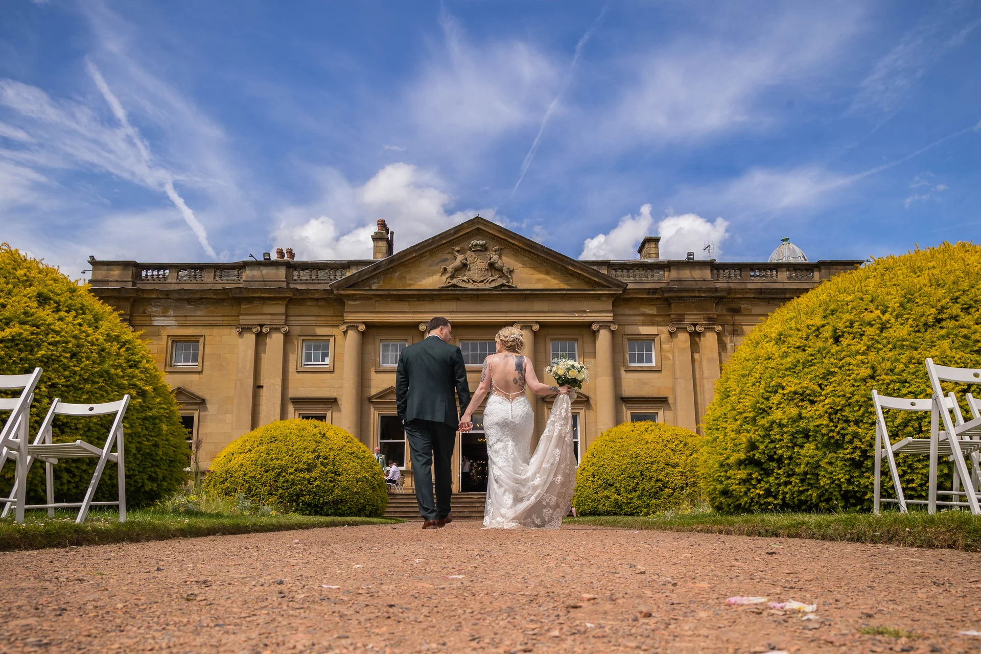 A newlywed couple walks hand in hand away from the camera along a gravel path toward a grand stately home under a bright blue sky.