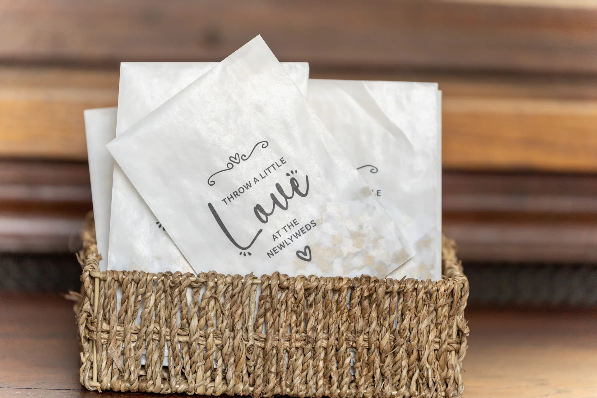 A woven wicker basket holds white paper confetti bags printed with the phrase throw a little love at the newlyweds.