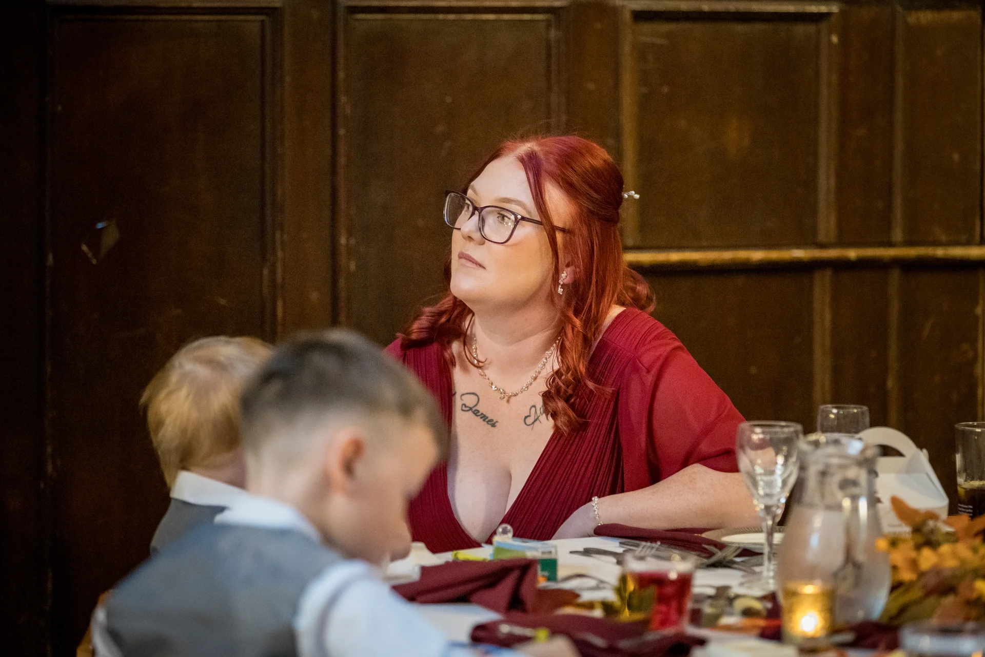 A guest with red hair and glasses looks attentively across the wedding breakfast table while seated near children.