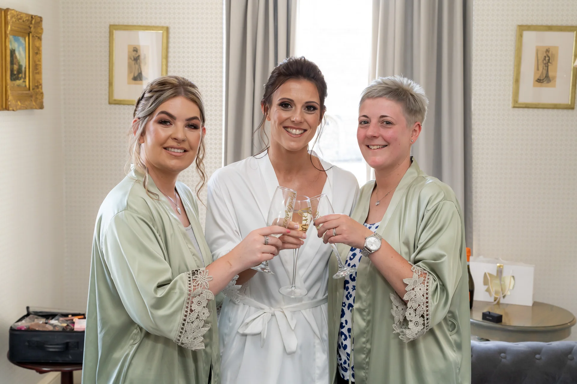 A smiling bride in a white robe toasts with champagne alongside two bridesmaids wearing matching sage green robes with lace trim.