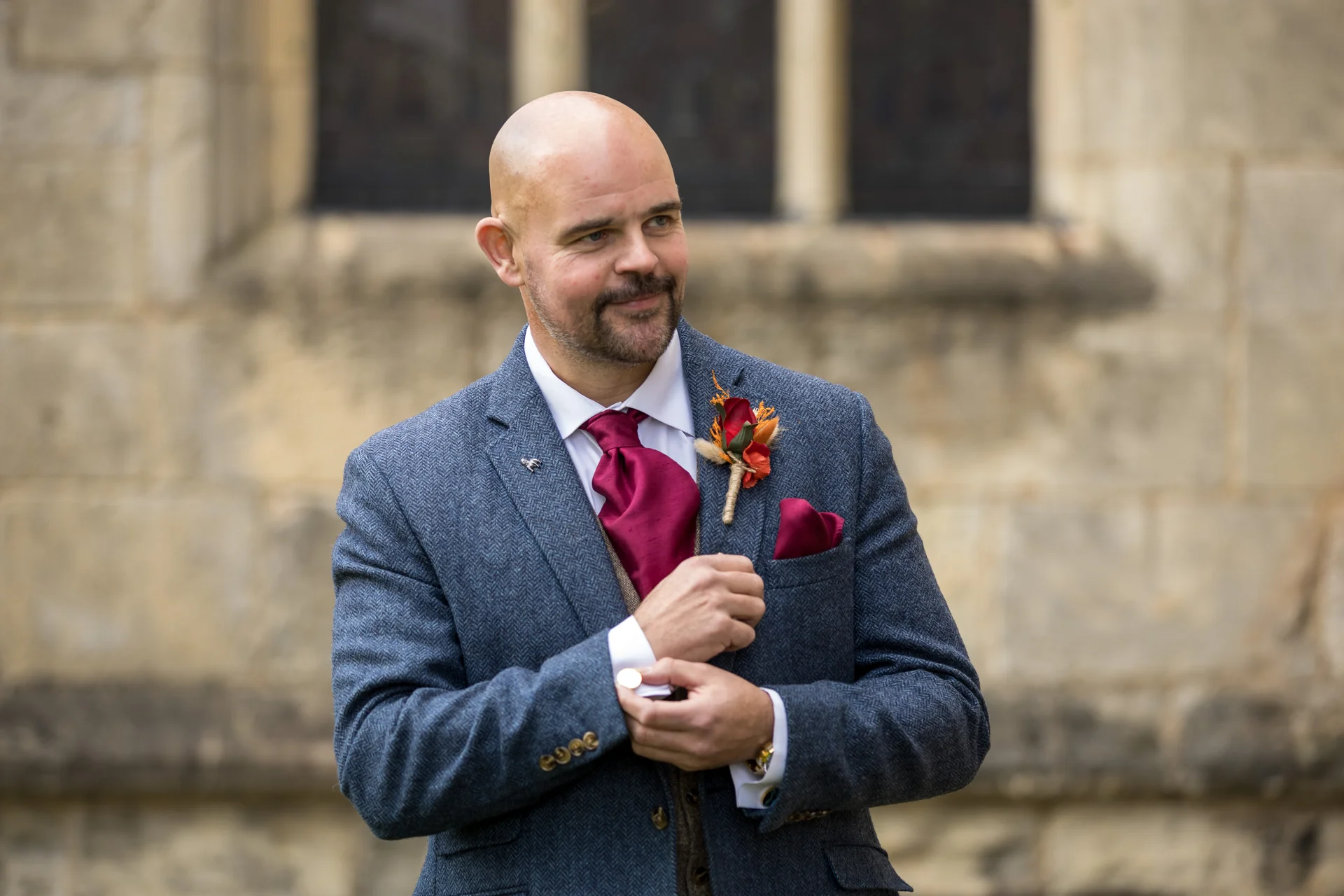 A smiling groom adjusts his cufflink while wearing a blue tweed suit, a rich burgundy tie, and a matching buttonhole outdoors against a stone background.