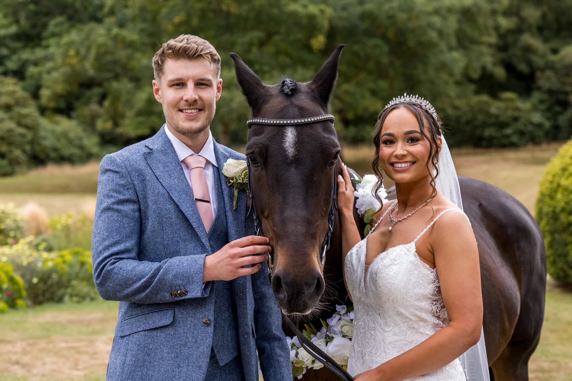 A smiling bride and groom pose outdoors next to a dark horse adorned with white floral decorations, set against a lush green background.