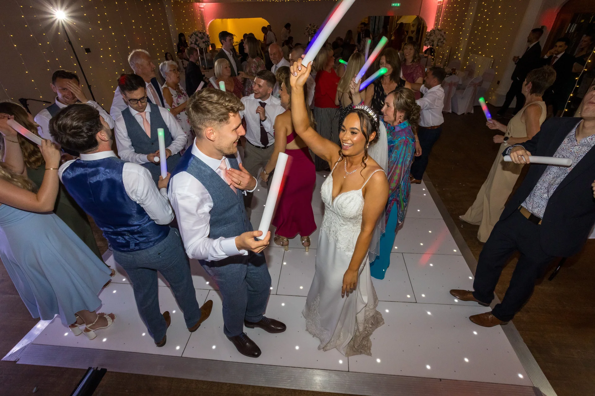 The bride and groom celebrate on a lit dance floor surrounded by joyful guests waving glowing foam sticks during the evening reception.