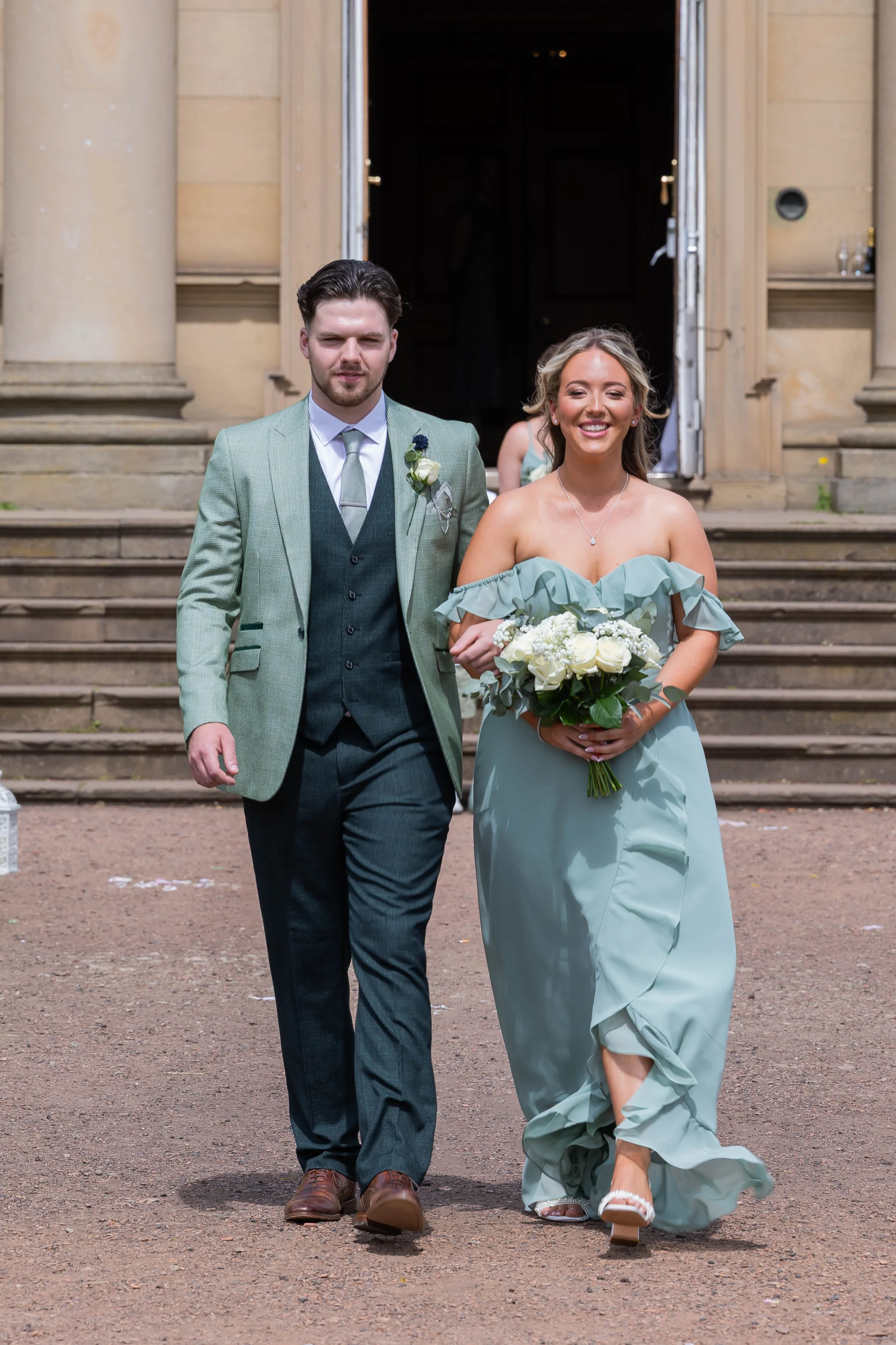 A smiling bride and groom walk hand in hand down stone steps outside a grand country house, with the groom in a green suit and the bride wearing an off-the-shoulder sage gown holding a white rose bouquet.