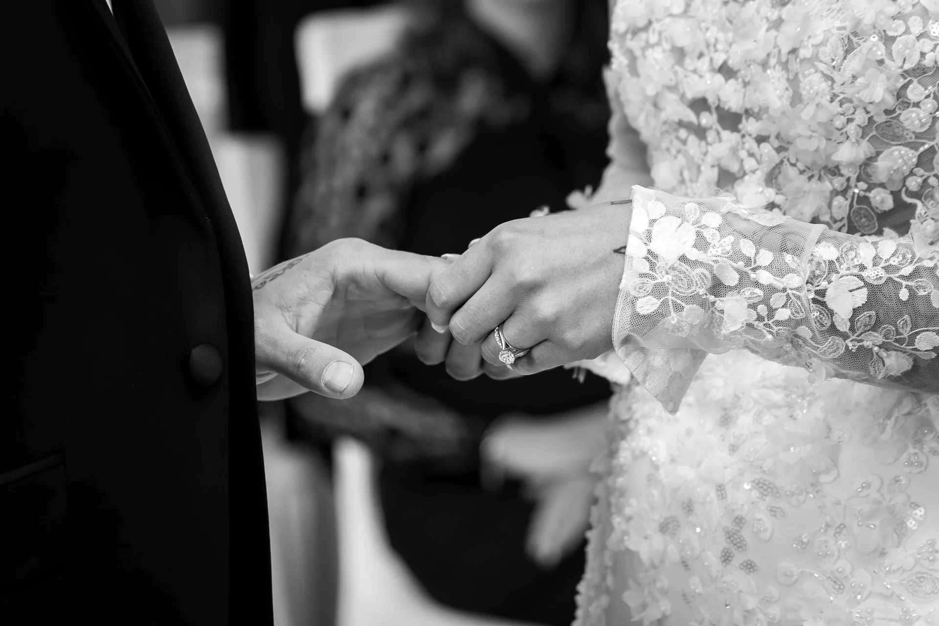 Close-up black and white photograph of a bride and groom exchanging rings during their wedding ceremony, showcasing the intricate lace sleeve of the bride's gown and her diamond engagement ring.