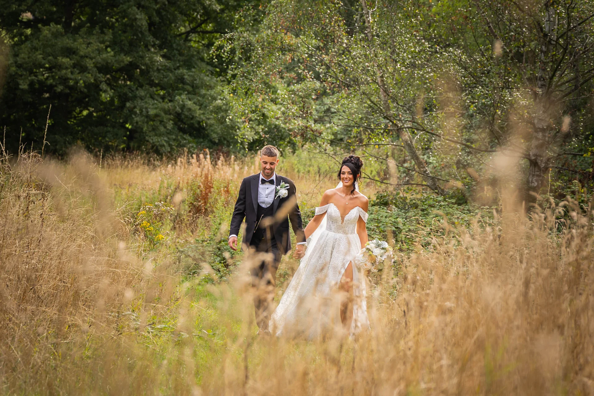 A newlywed couple happily walk hand-in-hand through a sunlit wildflower meadow and woodland setting, framed by tall golden grass in the foreground.