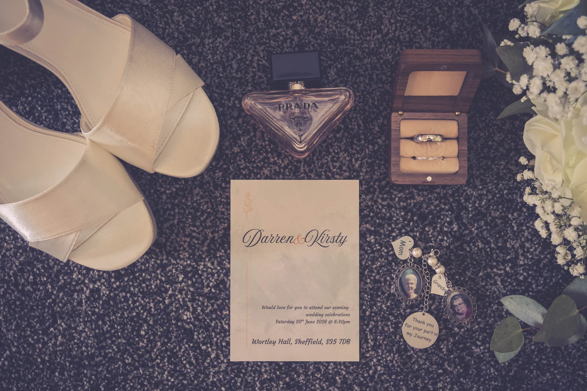 An elegant bridal flat lay showcases satin wedding shoes, a Prada perfume bottle, wedding rings in a wooden box, a custom invitation suite, and memorial charms.