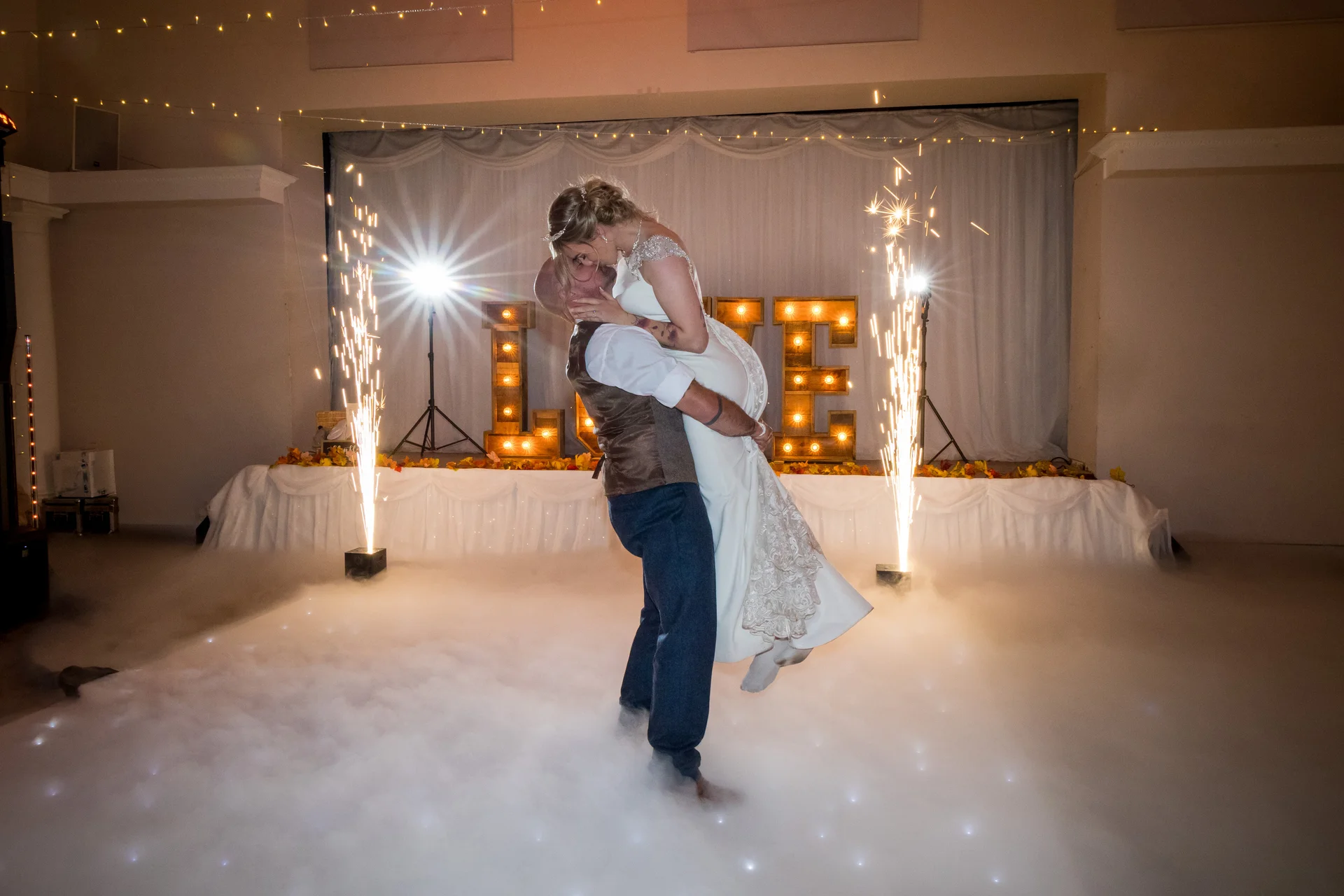 The groom lifts the bride into the air during their first dance on a smoke-filled dance floor, framed by indoor cold spark fountains and illuminated marquee letters.