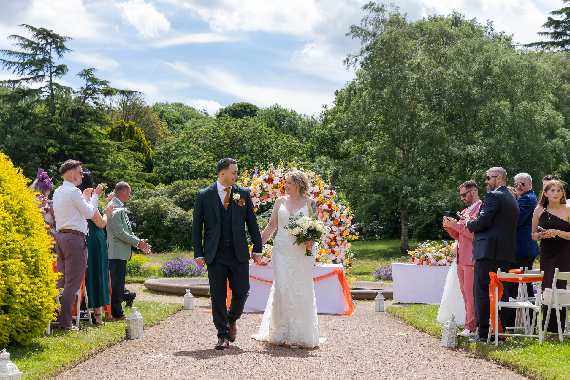 Newlyweds walk hand in hand down a gravel path after an outdoor garden ceremony, passing applauding guests and a vibrant floral arch.