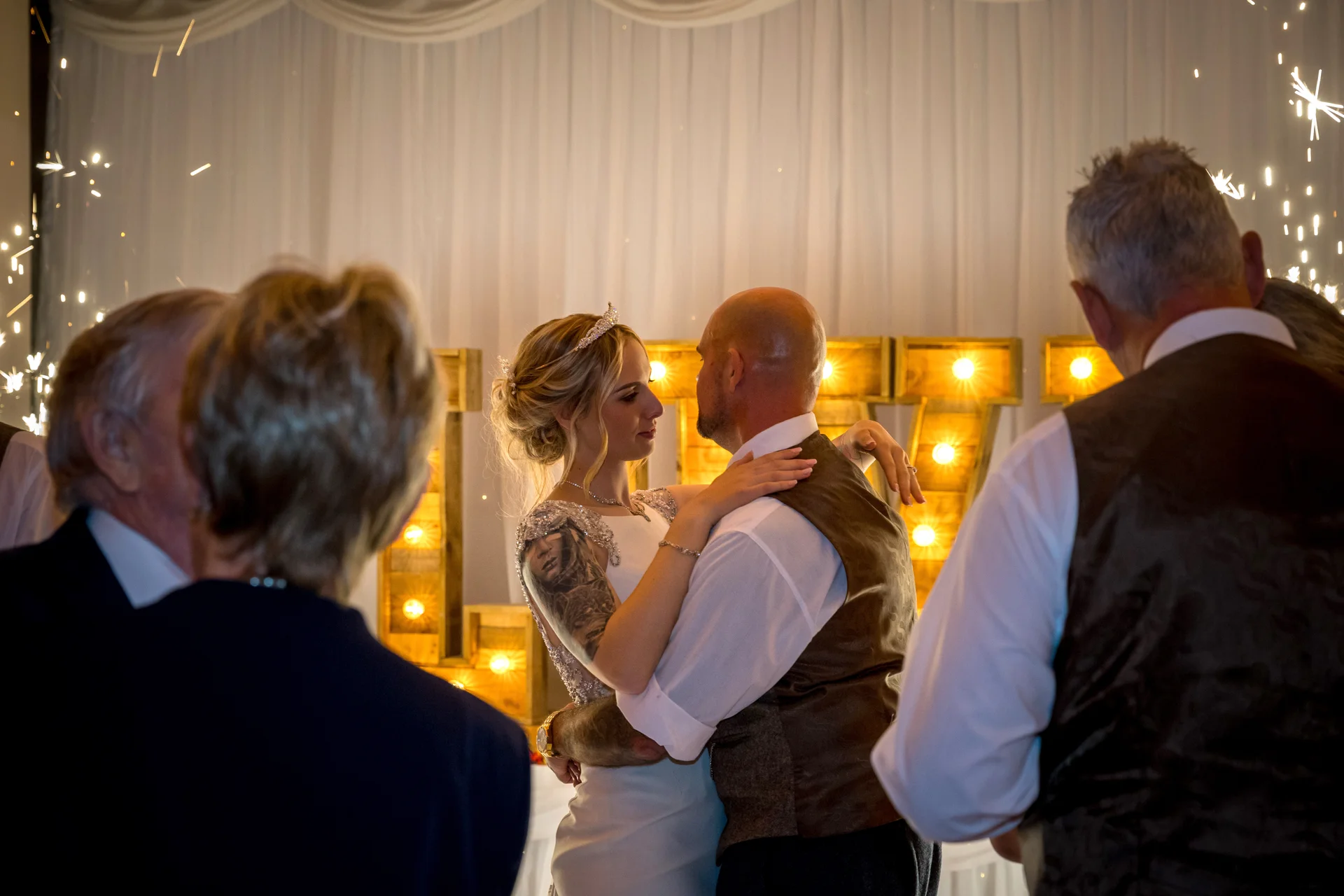 A newlywed couple share their first dance as guests look on, framed by illuminated letters and sparkling indoor fireworks.