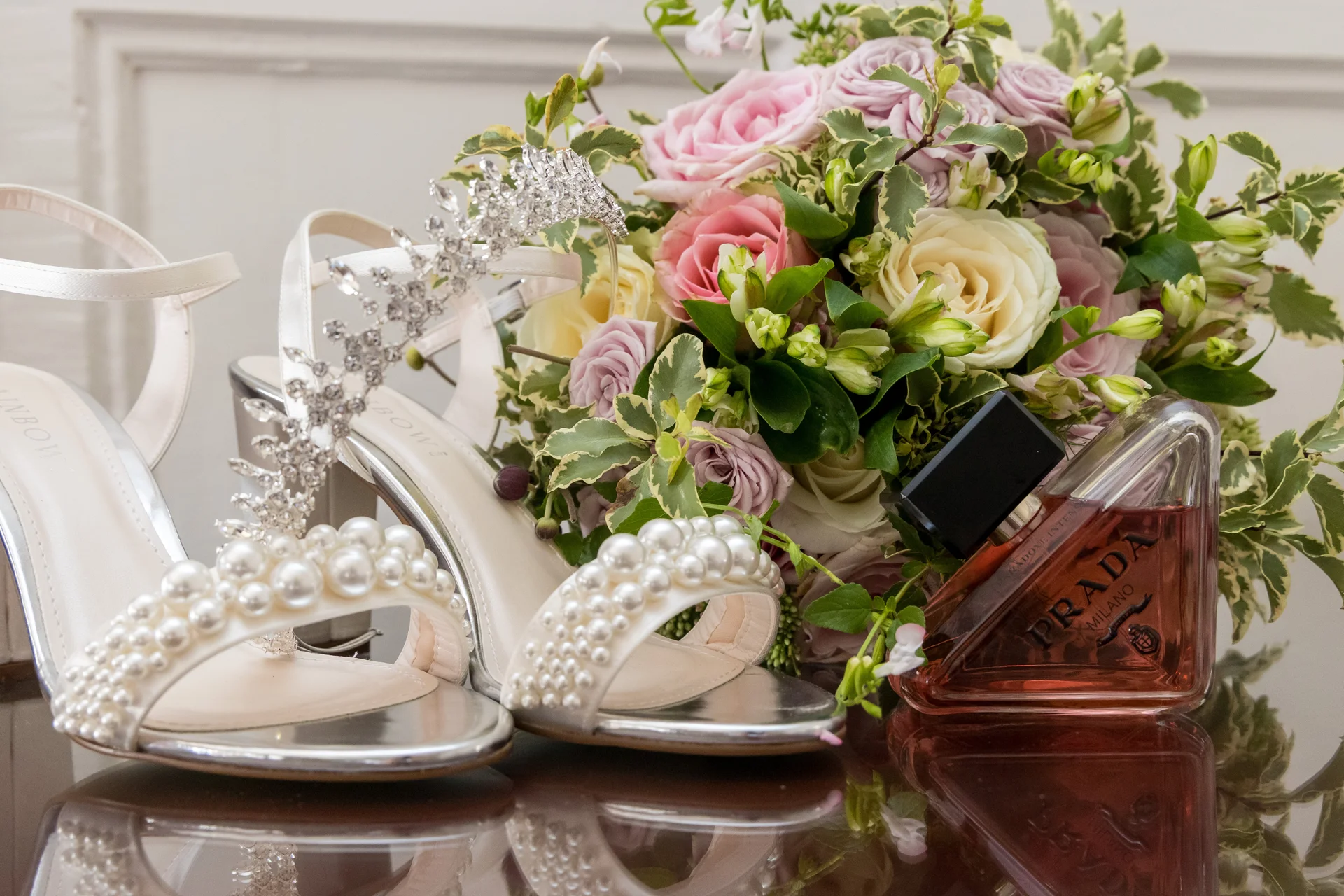 A bridal flat lay featuring pearl-adorned satin heels, a crystal hairpiece, a lush bouquet of pink and white roses, and a designer perfume bottle resting on a reflective surface.