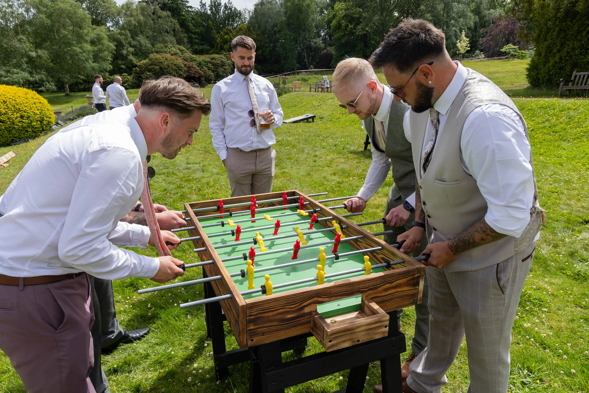 A group of groomsmen play table football on the lawn during a sunny outdoor wedding reception.