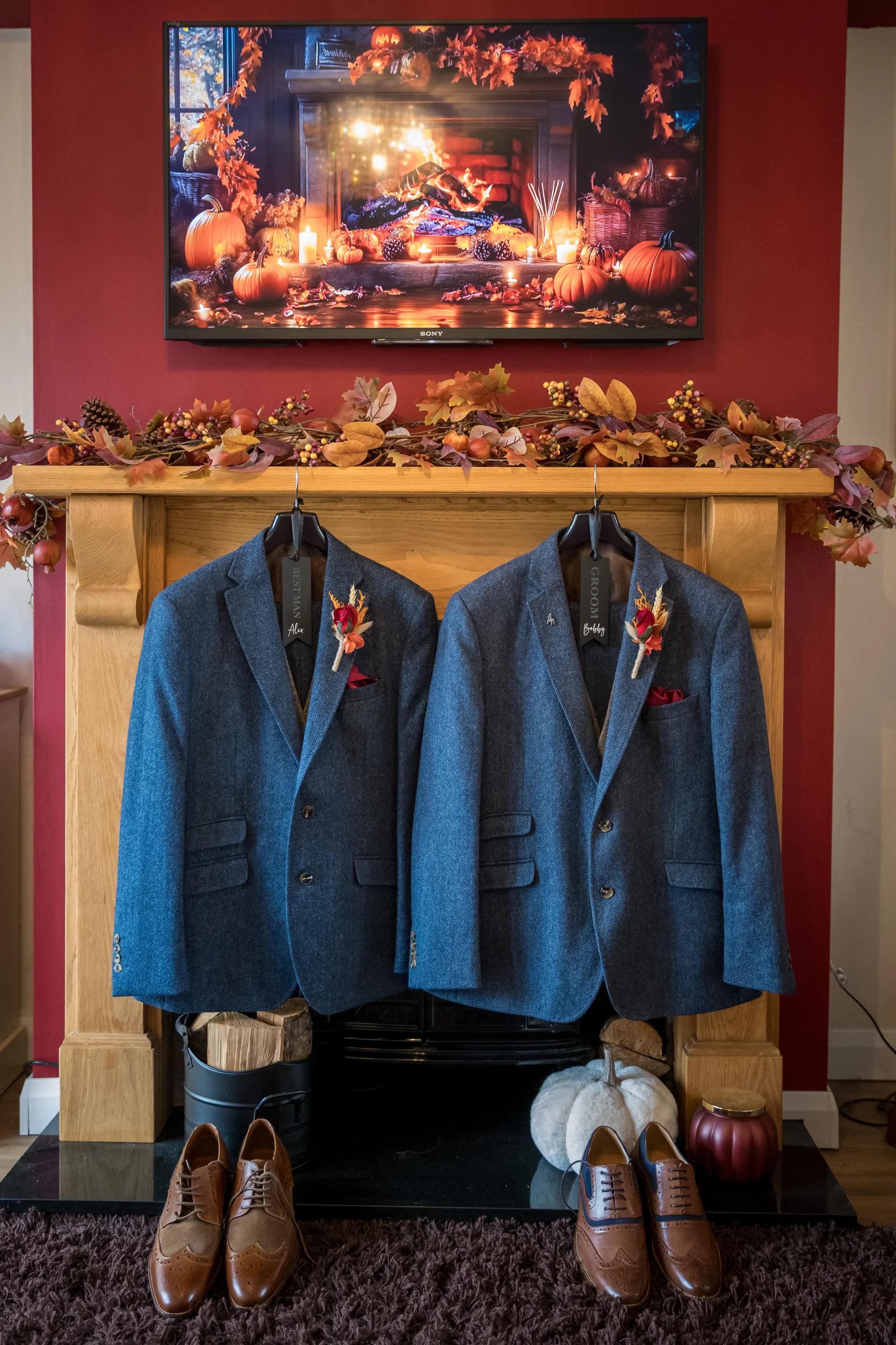 Two blue tweed groom and best man suits hang neatly from a wooden mantelpiece decorated with autumn foliage, positioned above a hearth with matching leather brogues and seasonal decor.
