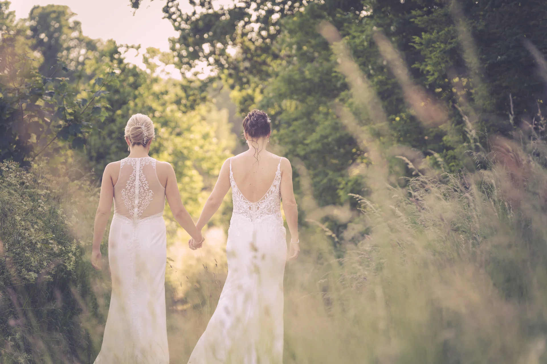 Two brides in elegant white wedding dresses walk hand in hand through a lush woodland path framed by tall grass and sun-dappled trees.