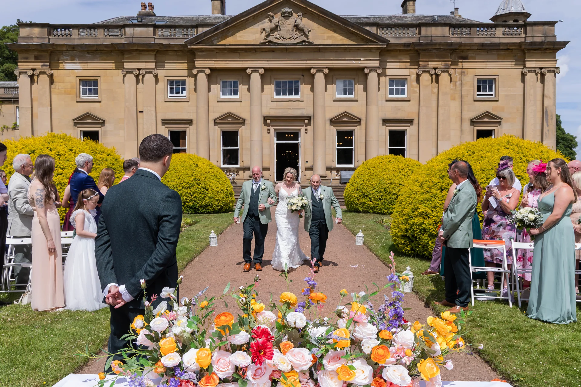 The bride walks down an outdoor aisle flanked by family members and guests towards the waiting groom in front of a grand country house.