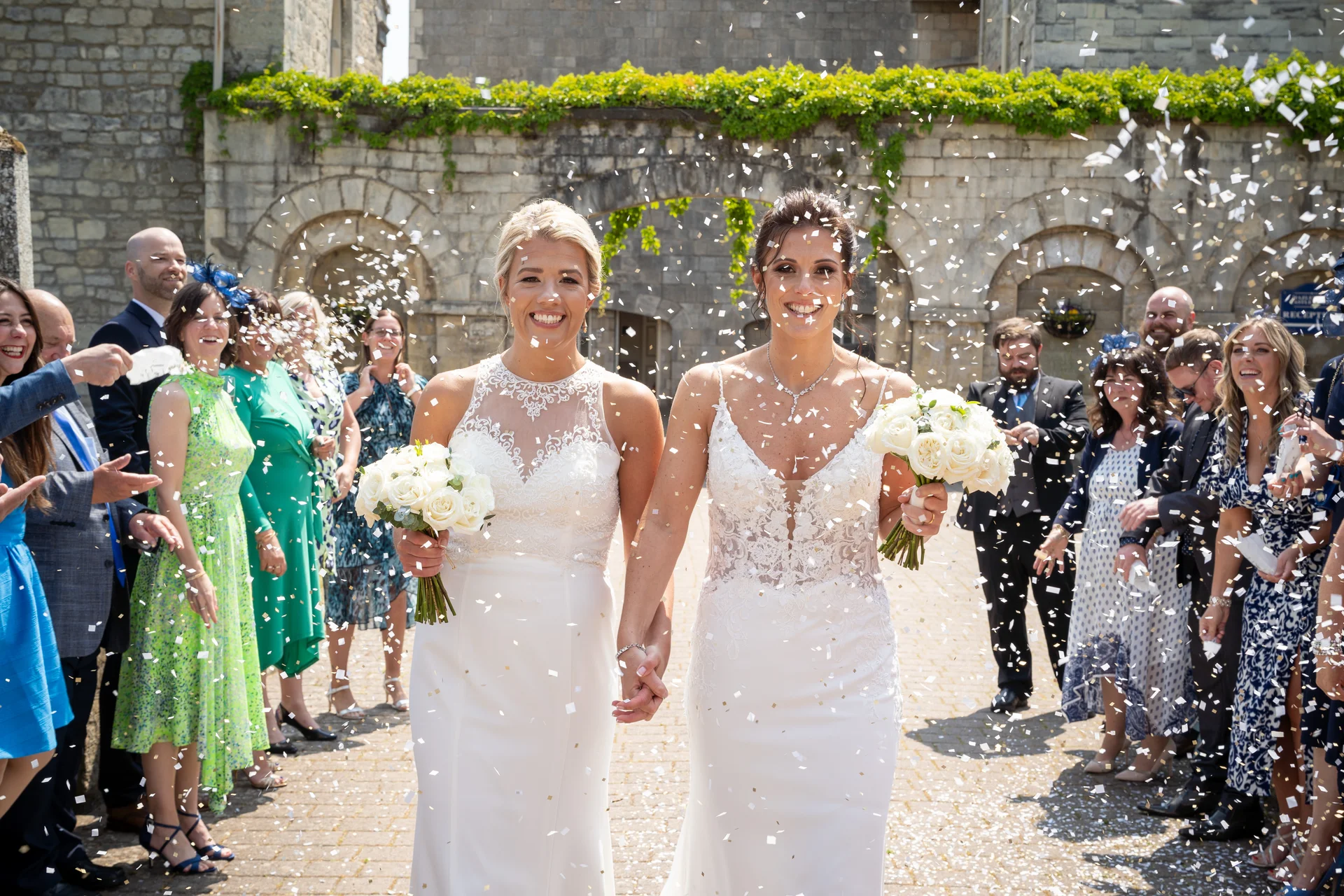 Two smiling brides walk hand-in-hand through a shower of white confetti, holding bouquets of white roses outside a historic stone castle courtyard.