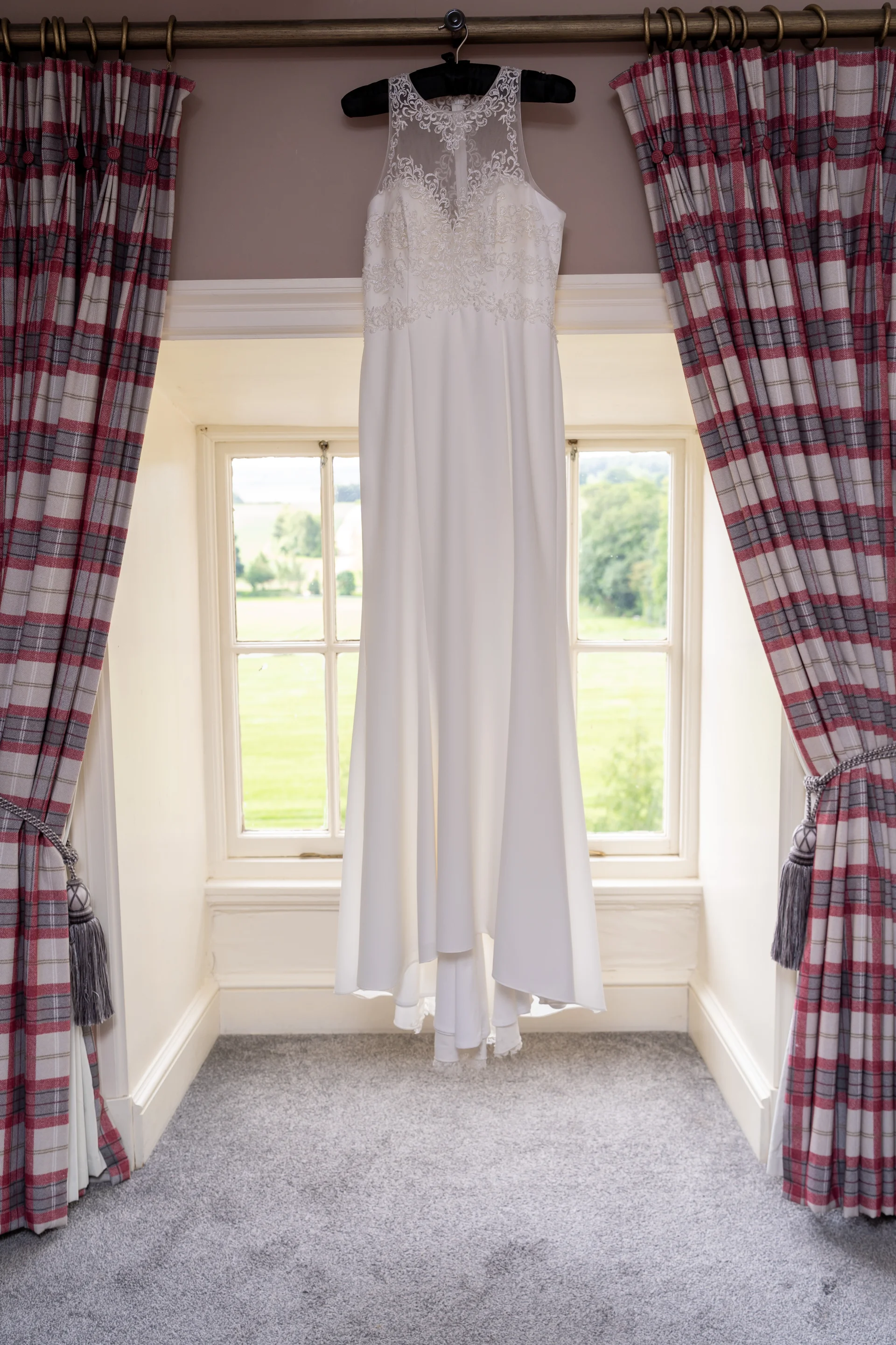 An elegant white wedding dress with an illusion lace neckline hangs gracefully in front of a bright window framed by patterned curtains.