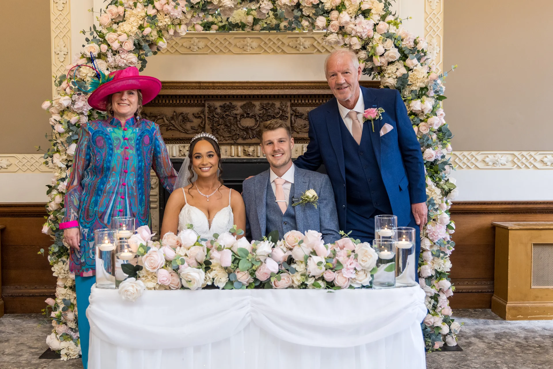The newly married couple sits together at a draped top table flanked by family members under a large floral arch adorned with pink roses.