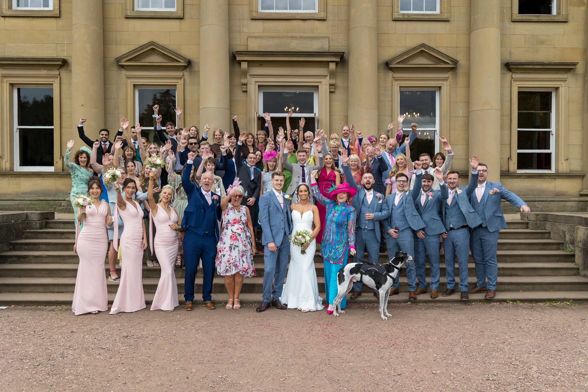 The wedding party and guests cheer and raise their arms in celebration on the stone steps outside a grand country house, alongside a dog on a leash.