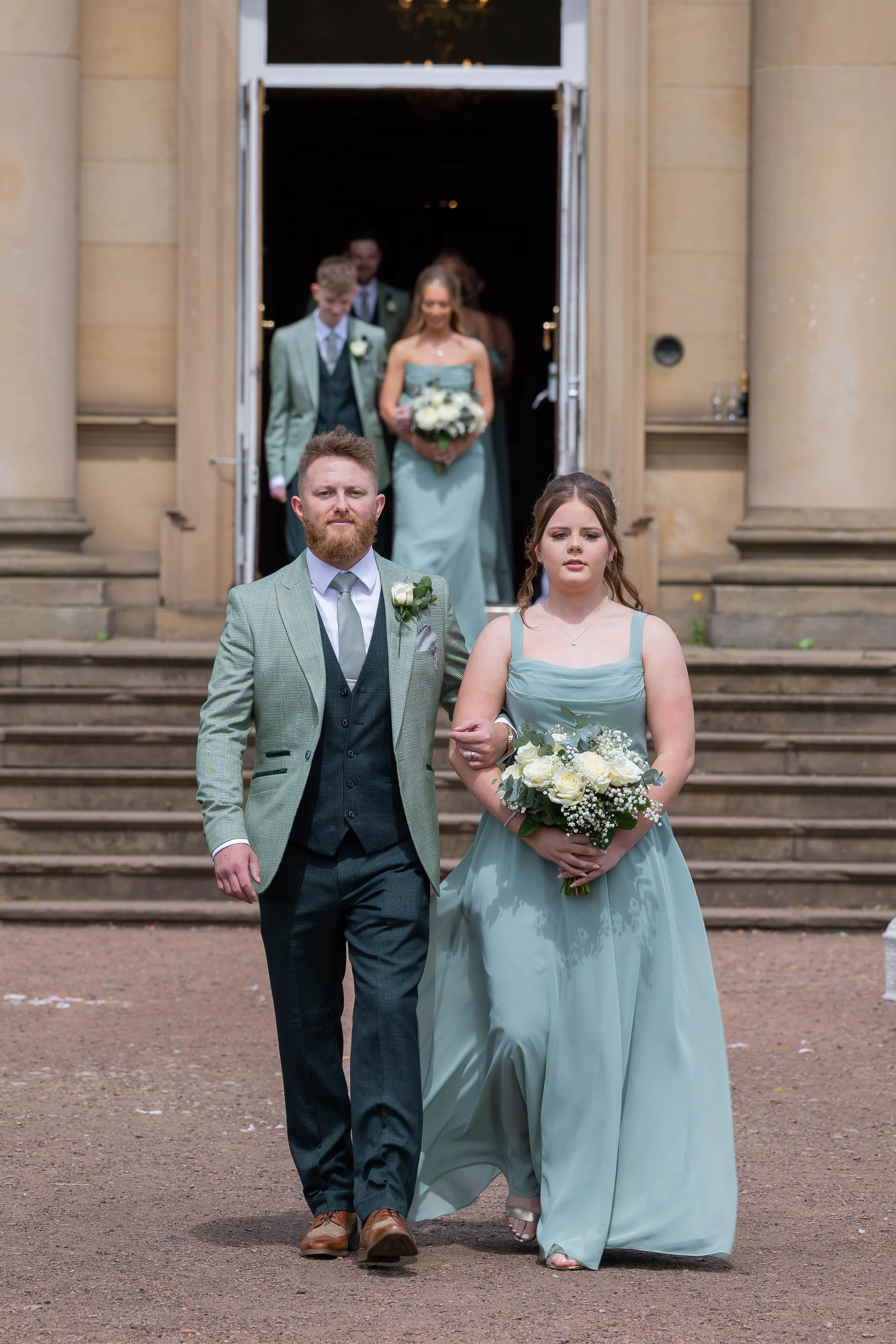 A groom in a sage green tweed suit and a bridesmaid in a sage dress walk arm-in-arm out of a grand country house doorway with wedding guests following behind.
