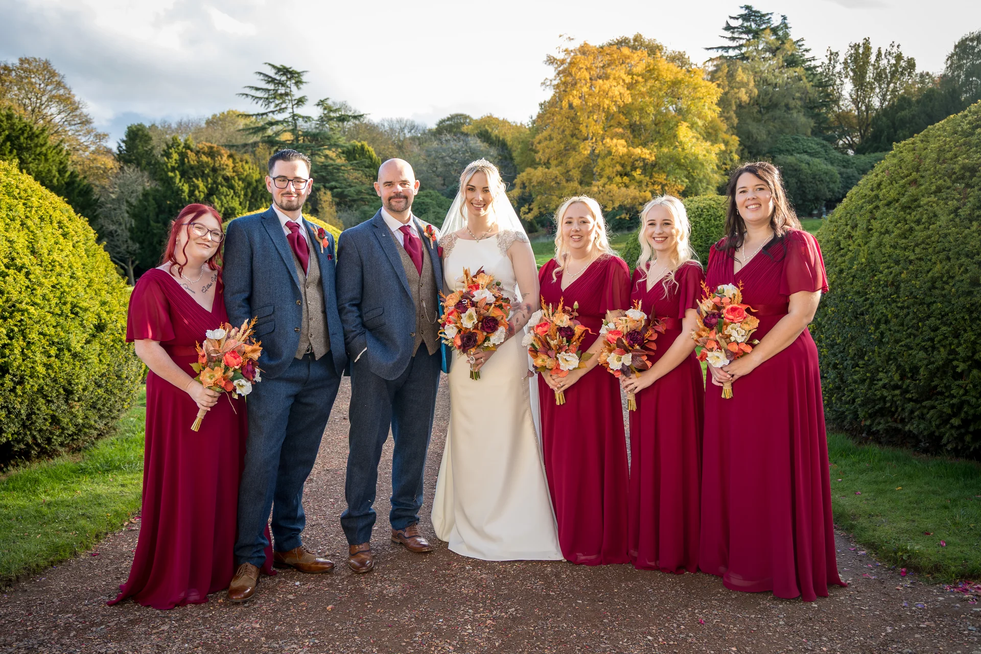 A newlywed couple poses on a gravel pathway in a manicured garden alongside their groomsmen in tweed suits and bridesmaids in long burgundy dresses holding vibrant autumn bouquets.