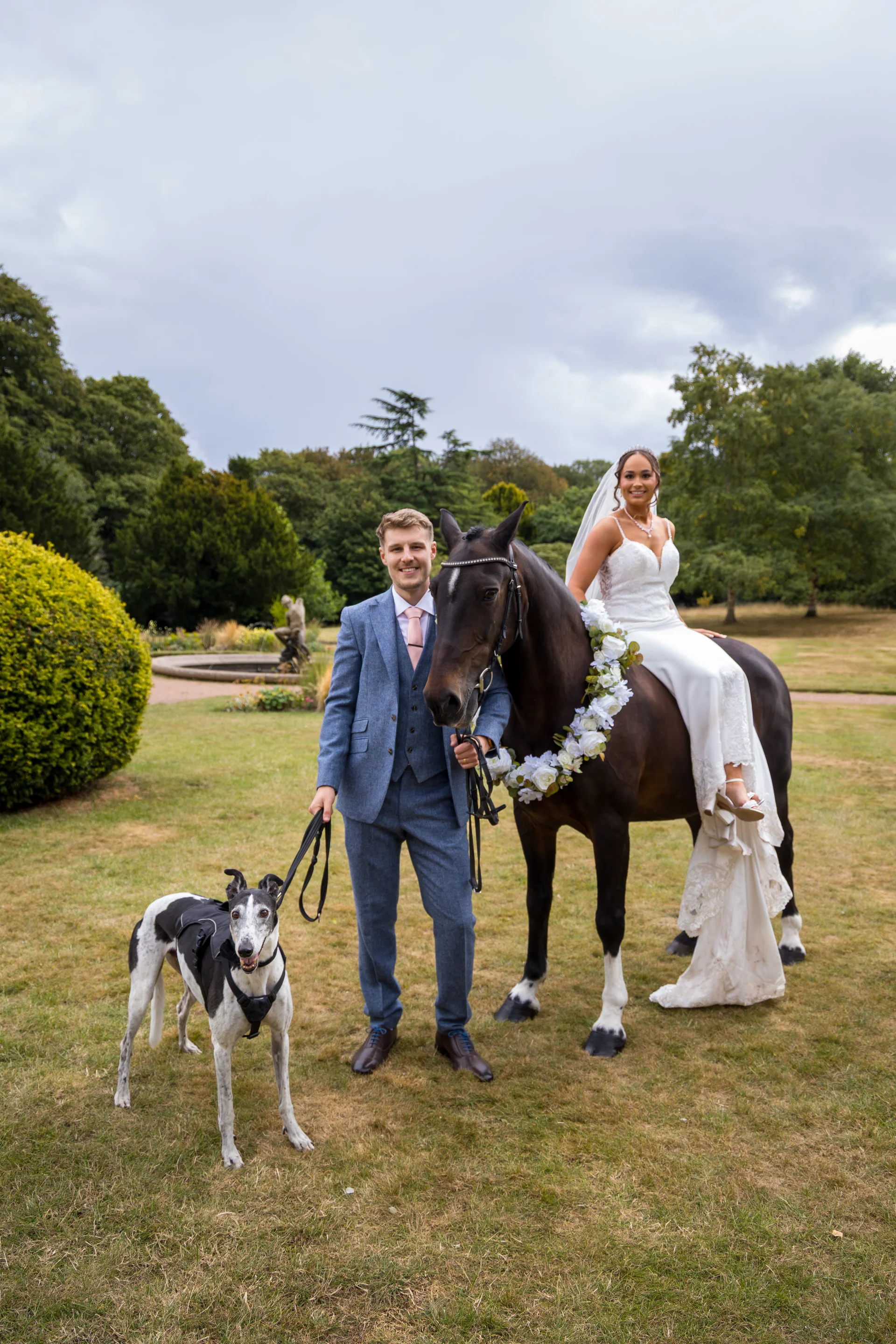 A newlywed couple pose outdoors in a country garden, with the groom standing holding a dog on a leash while the bride sits gracefully atop a horse adorned with a floral collar.