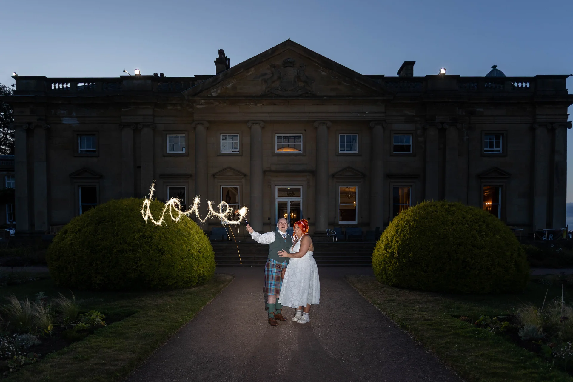 A newlywed couple poses in front of a grand illuminated stately home at dusk, while a sparkler writes the word love in the air.