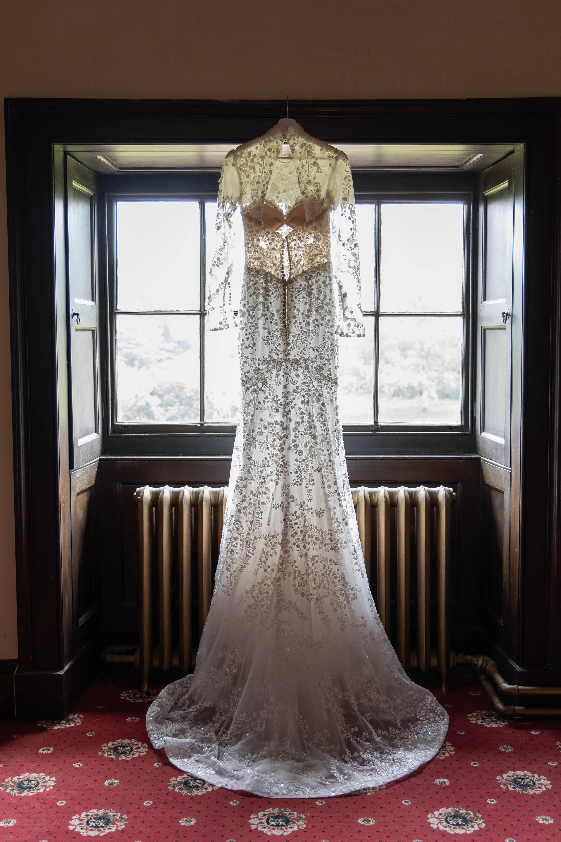 An intricate long-sleeve lace wedding dress hangs gracefully against a bright window inside a traditional room.