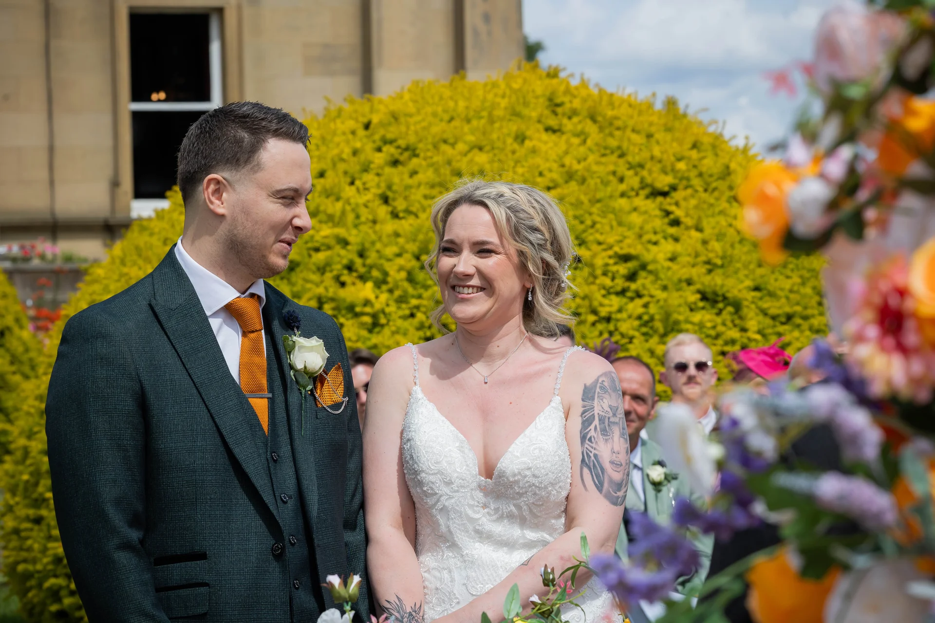 A smiling bride and groom stand together during an outdoor wedding ceremony, framed by vibrant orange and purple floral installations.
