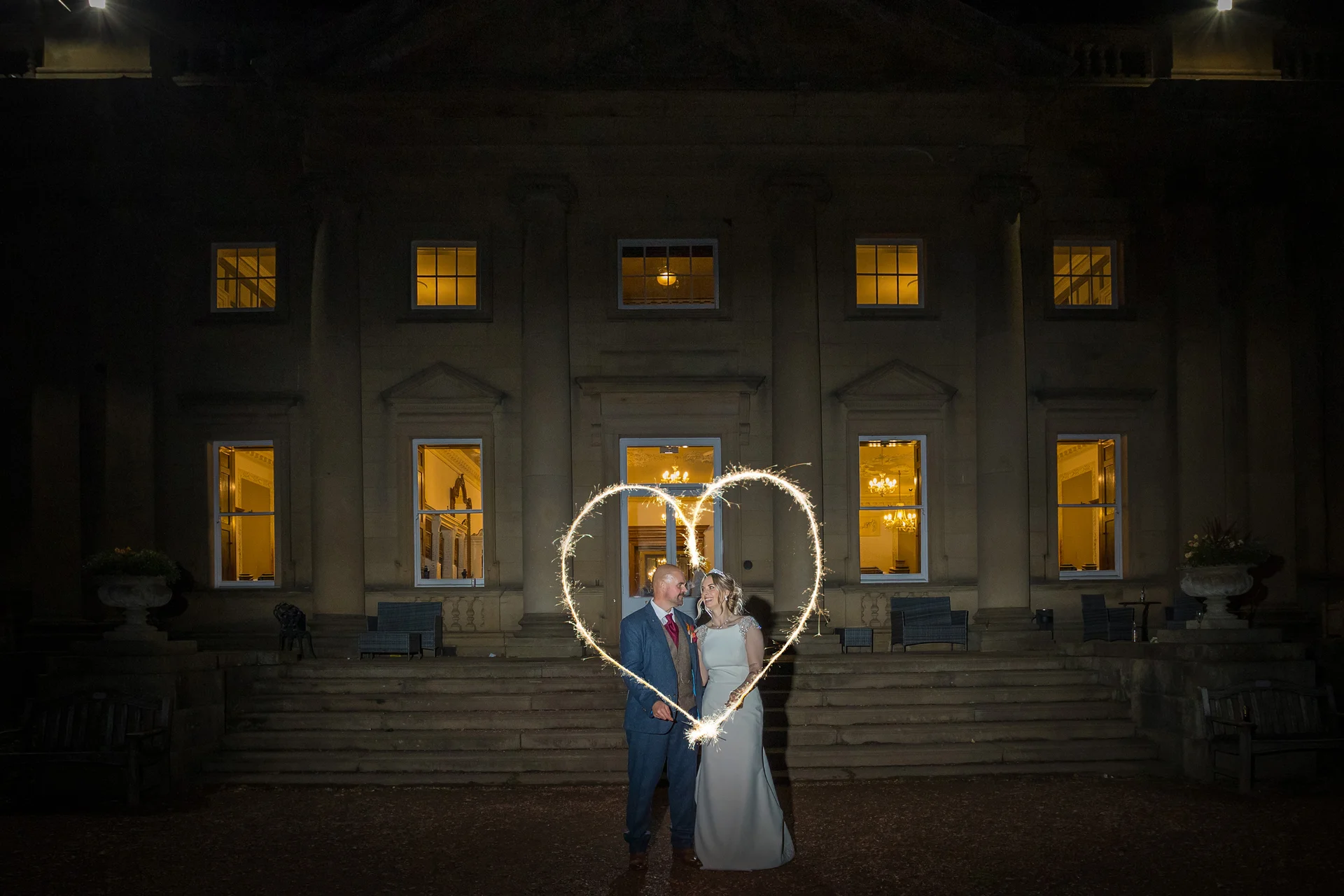 A newlywed couple stand smiling in front of a grand illuminated country house at night while a sparkler light painting creates a glowing heart shape around them.