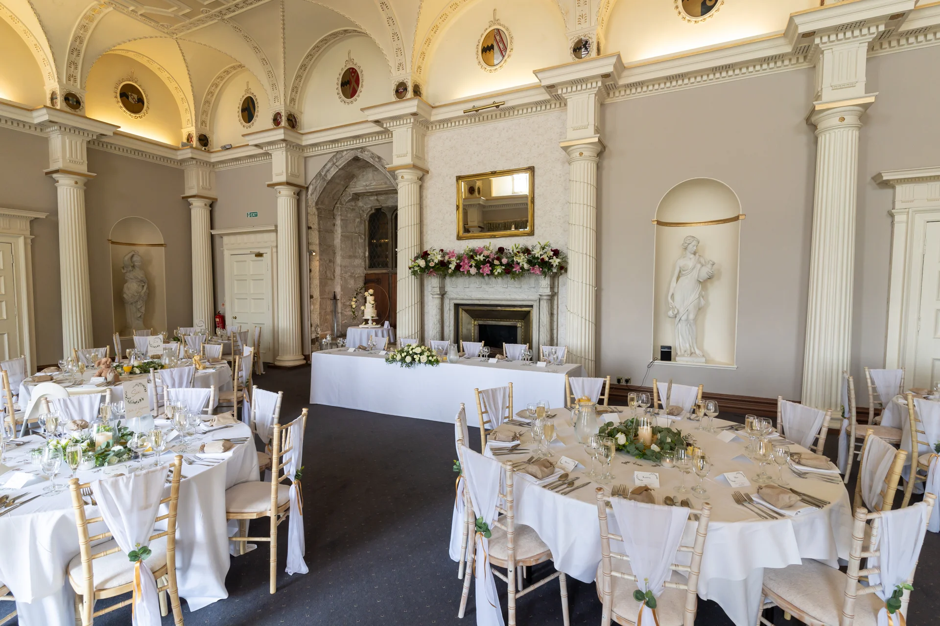 A grand stately home ballroom set up for a wedding breakfast, featuring round guest tables and a top table decorated with white linens and floral arrangements.