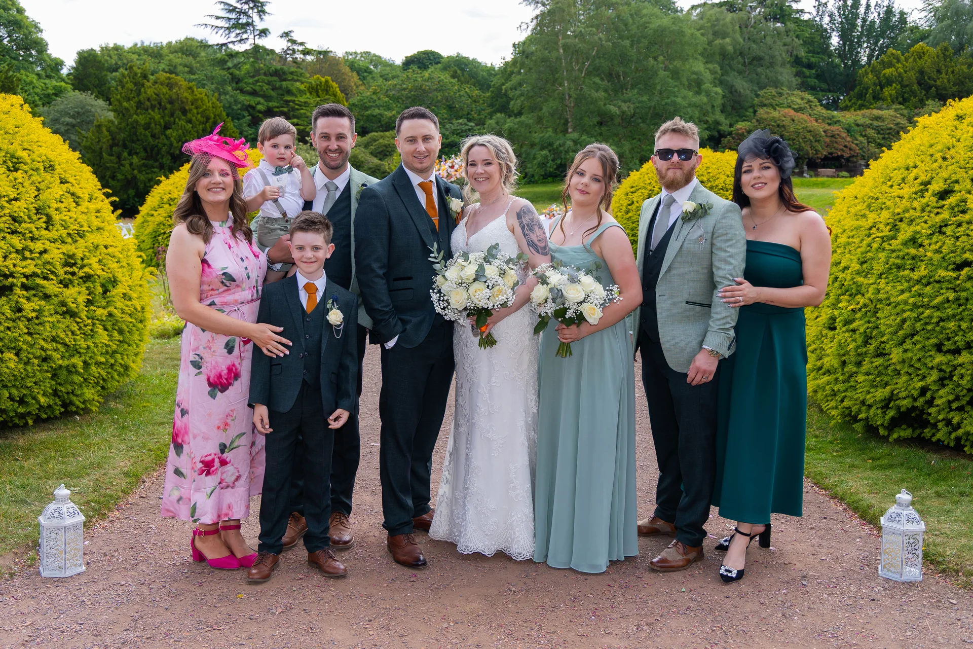 A large wedding party stands smiling together on a gravel garden path flanked by bright yellow bushes, featuring the bride in a lace gown and the groom in a dark suit.