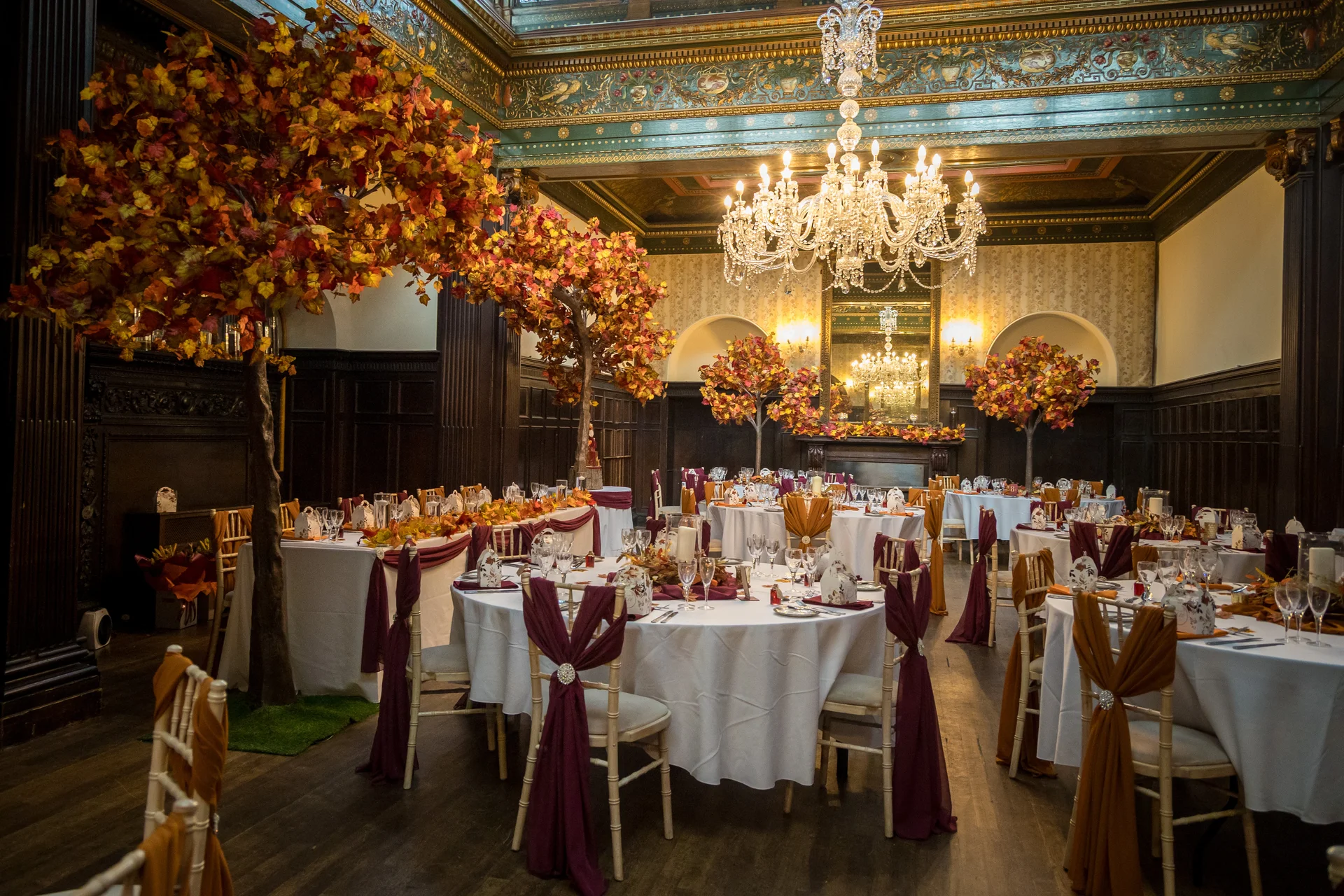 An ornate historic ballroom is set up for an autumn wedding reception with round tables decorated in white linens, burgundy chair sashes, and striking artificial autumn trees with vibrant orange and red foliage.