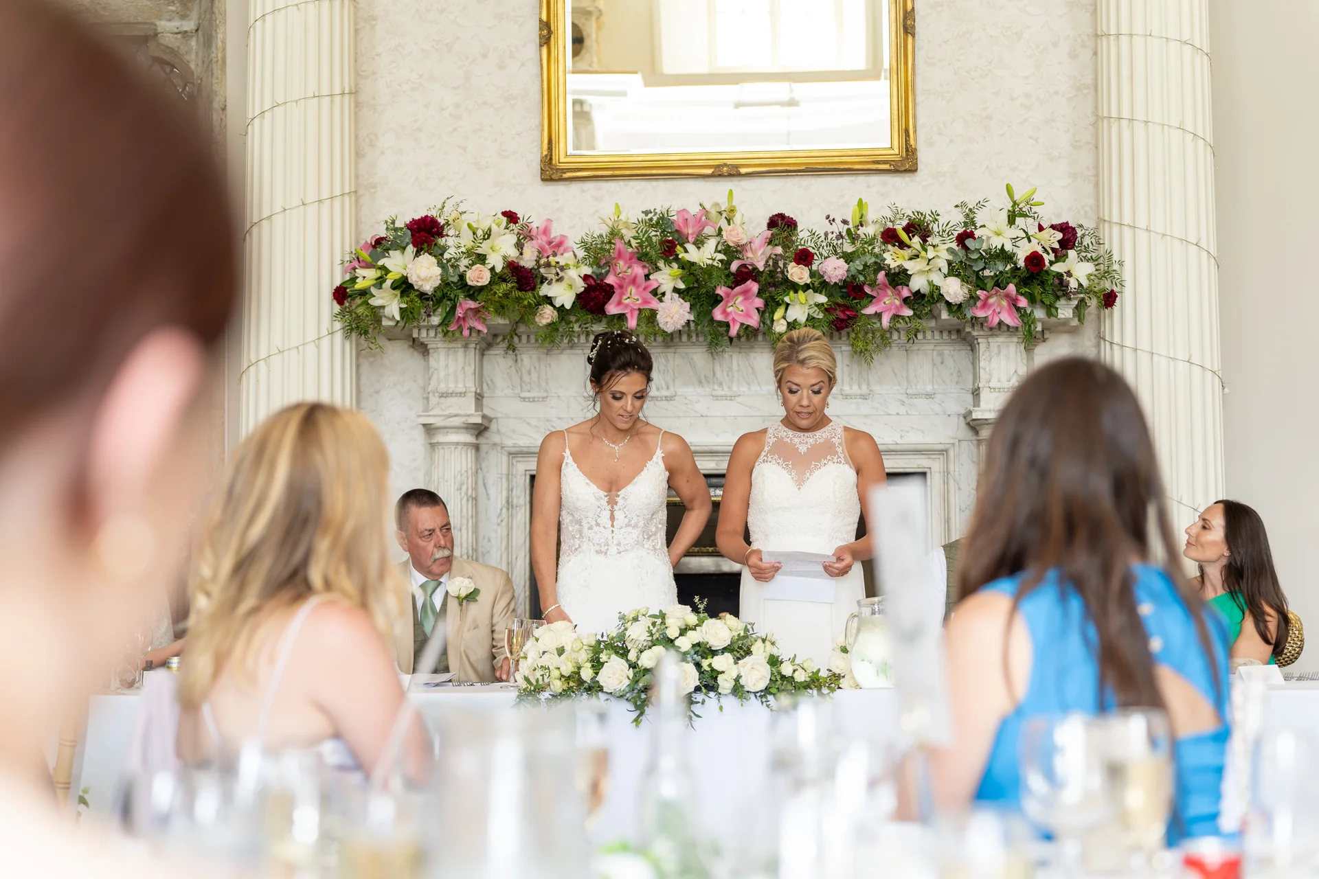 Two brides stand together at the top table delivering wedding speeches in front of a grand fireplace adorned with a vibrant floral garland.